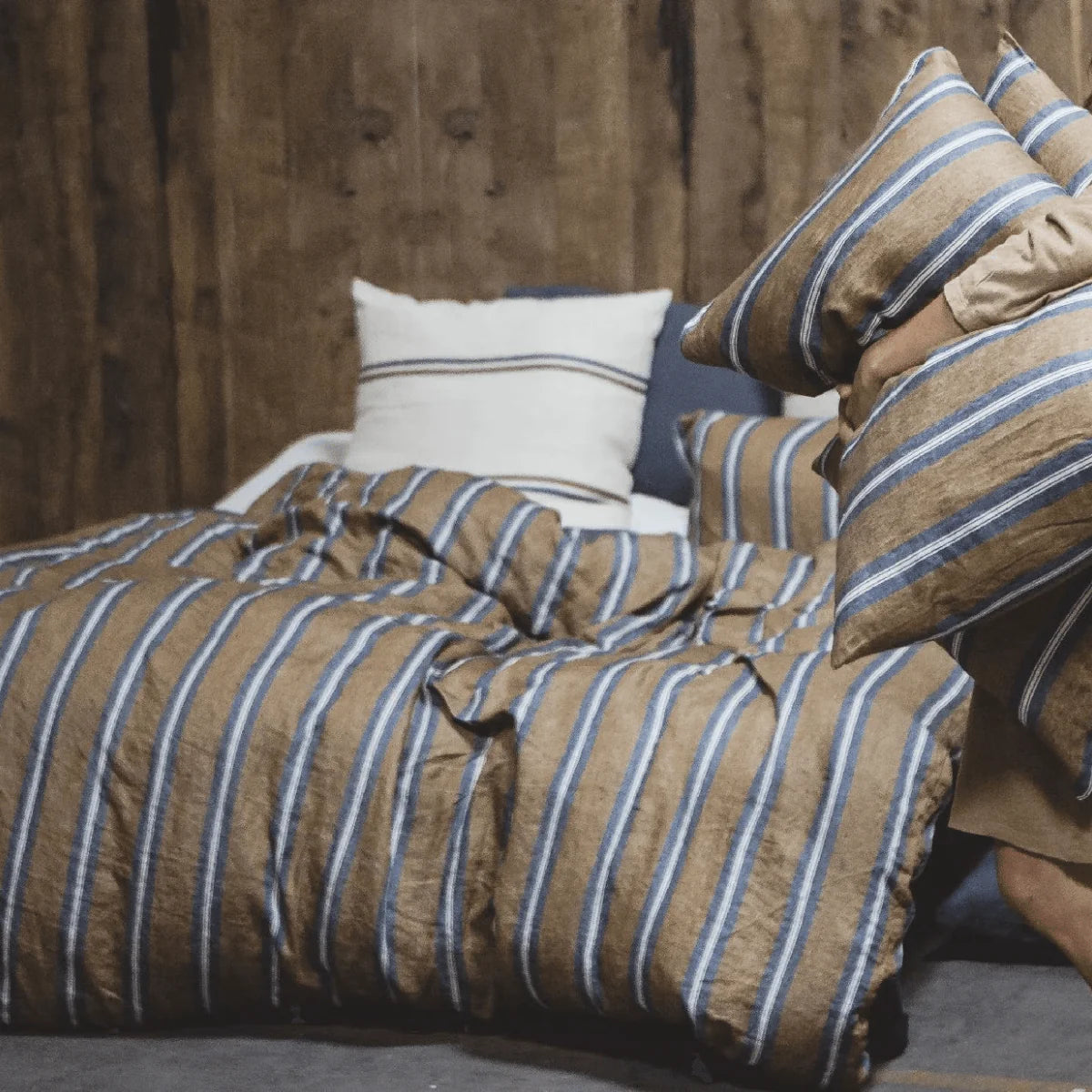 A person sits on a bed with brown and blue striped bedding, holding two matching pillows. Wooden paneling and the Libeco Home Salem Duvet Cover Stripe Twin create a cozy atmosphere, promising a comfortable sleep.