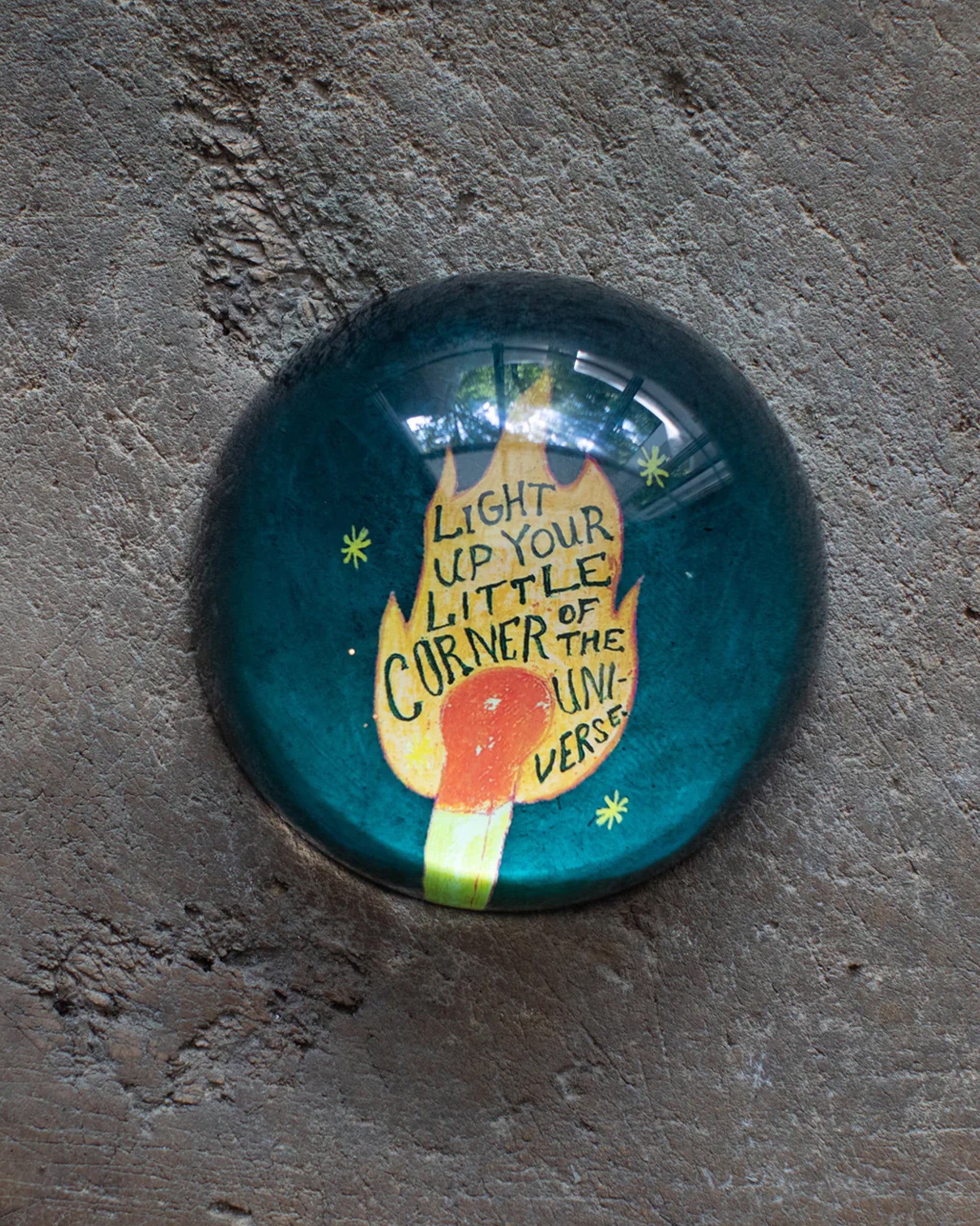 The "Light up" Paperweight by Sugarboo Designs features a lit match illustration and the quote, "Light up your little corner of the universe," surrounded by yellow stars on a textured surface—a unique desk accessory.