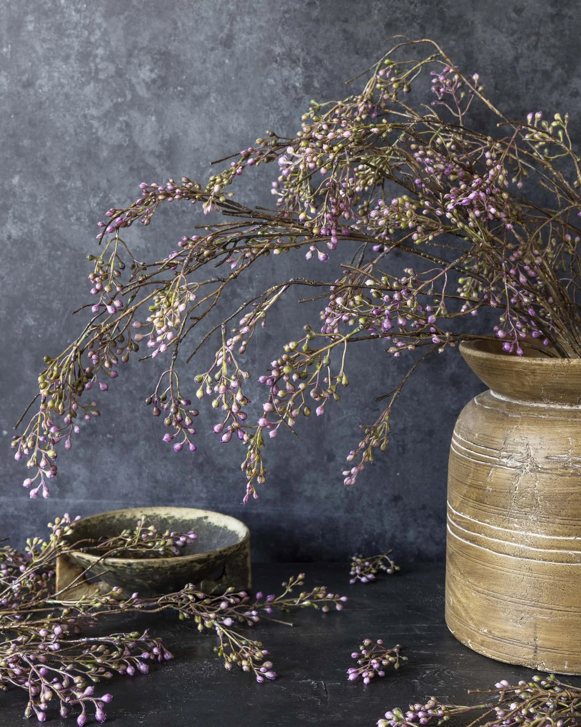 The Berry Branch Lilac by Abigail Ahern sits in a rustic ceramic vase, its purple buds and artificial berries creating a striking scene. Some branches and berries are scattered nearby on the dark surface with a small bowl, set against a textured gray backdrop.