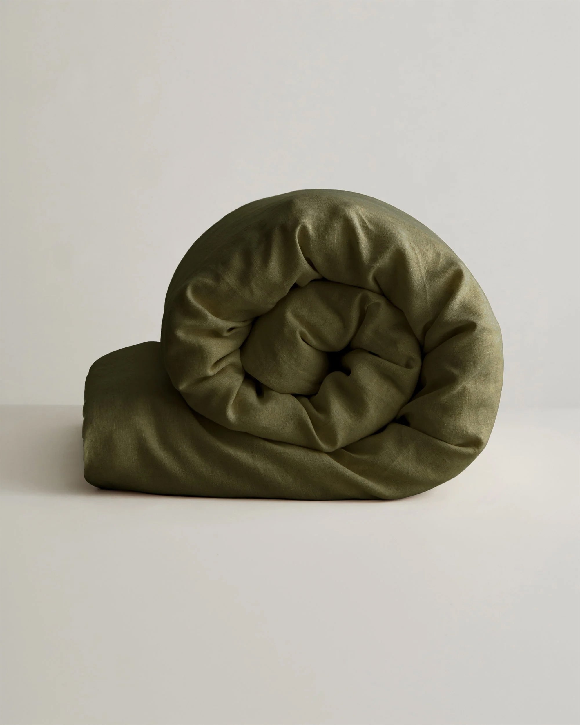 Green linen duvet cover by Cultiver Goods, rolled up on a pale surface with a minimalist neutral background.