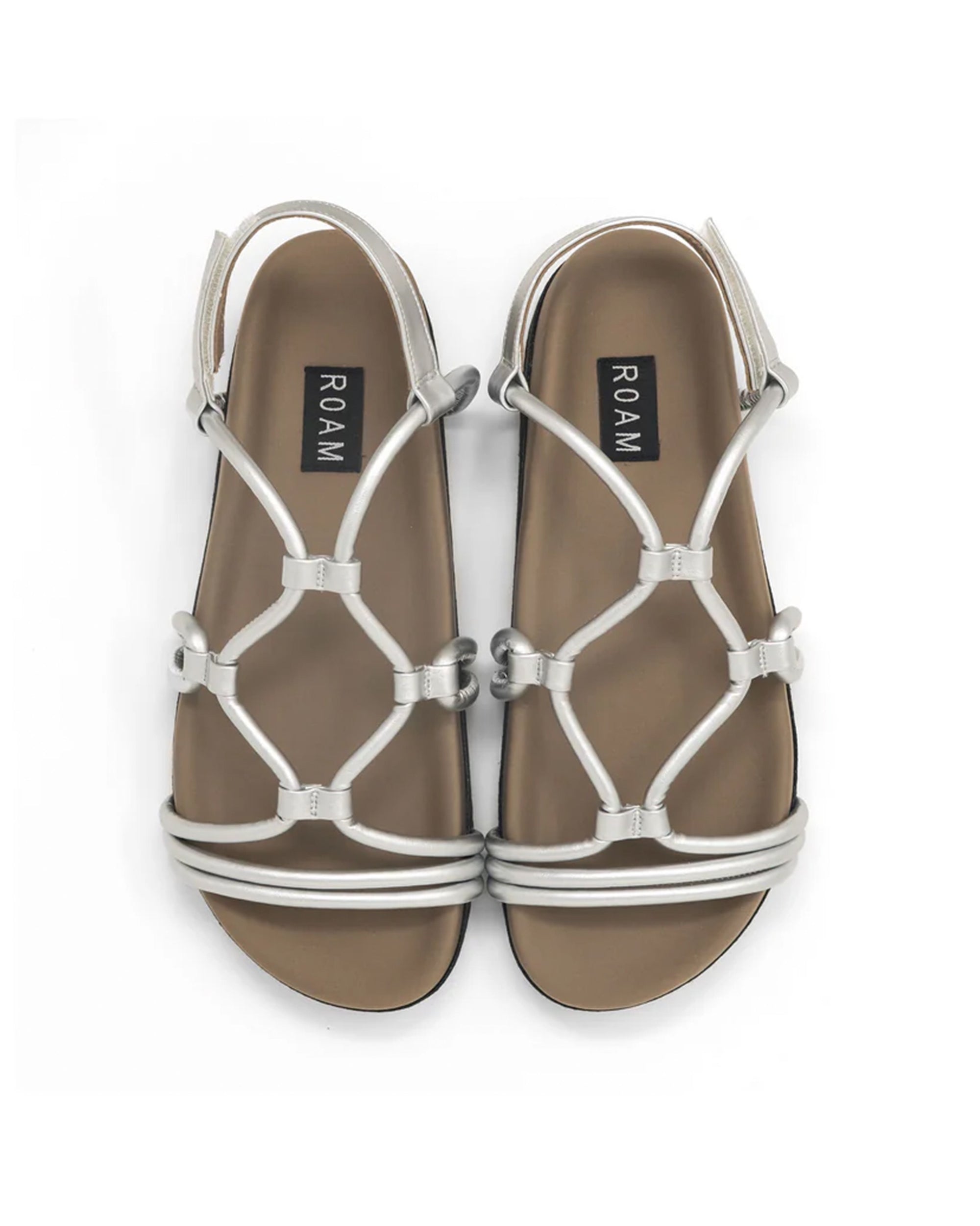 The Roam Linky Sandal features tan contoured footbeds and metallic silver vegan leather crisscross straps, with "ROAM" labels on the insoles. Shown from above on a white background.