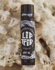 A black tube of Faire Lip Trip SPF lip balm sits on a textured beige surface resembling coral, with white decorative accents and bold text on the label emphasizing its sun protection benefits.