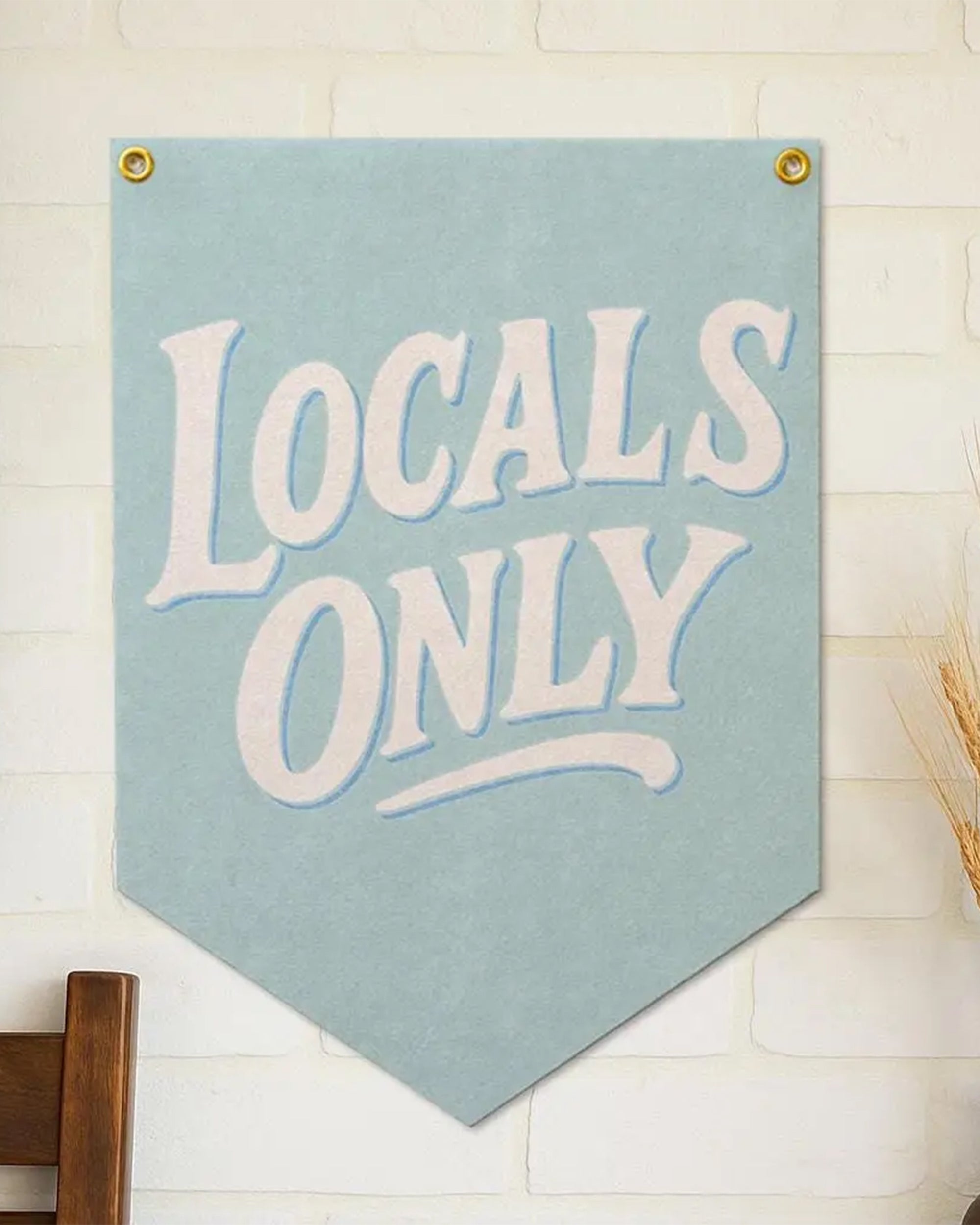 A light blue "Locals Only" Felt Flag by Faire hangs on a white brick wall above a wooden chair, adding vintage coastal banner style to your beach wall decor.