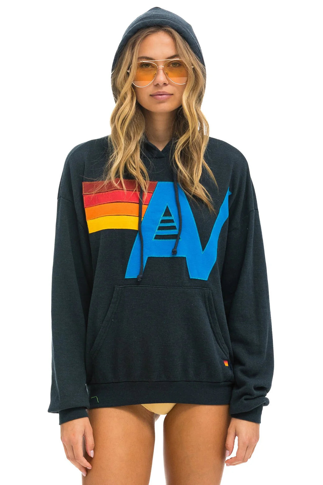 A person is wearing the logo stitch pullover hoodie from Aviator Nation, featuring colorful red, orange, yellow, and blue designs. They have long hair and are sporting yellow-tinted sunglasses while standing against a white background.