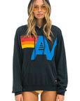 A person is wearing the logo stitch pullover hoodie from Aviator Nation, featuring colorful red, orange, yellow, and blue designs. They have long hair and are sporting yellow-tinted sunglasses while standing against a white background.