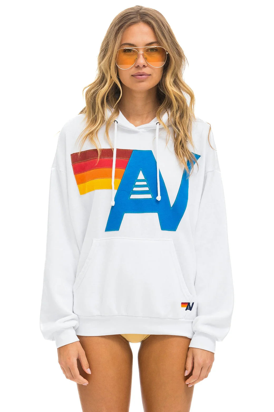 A woman with long hair is wearing orange-tinted sunglasses and the Aviator Nation logo stitch pullover hoodie in a relaxed fit, which showcases a bold, colorful graphic design. She stands against a plain white background.