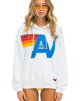 A woman with long hair is wearing orange-tinted sunglasses and the Aviator Nation logo stitch pullover hoodie in a relaxed fit, which showcases a bold, colorful graphic design. She stands against a plain white background.