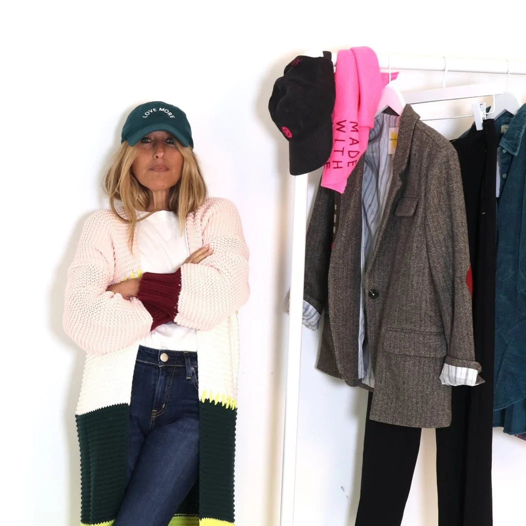 A person with long hair stands with arms crossed, wearing a white and green cardigan, jeans, and the Kerri Rosenthal Love More Hat in emerald green cotton. Behind them is a clothing rack adorned with various items, including a grey blazer, a pink scarf, and a black hat.