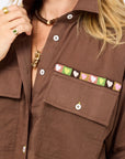 A person wears the Kerri Rosenthal Tia Love Brigade Shirt, a relaxed brown button-down with colorful embroidered hearts above the left pocket, styled with gold jewelry and a green ring.