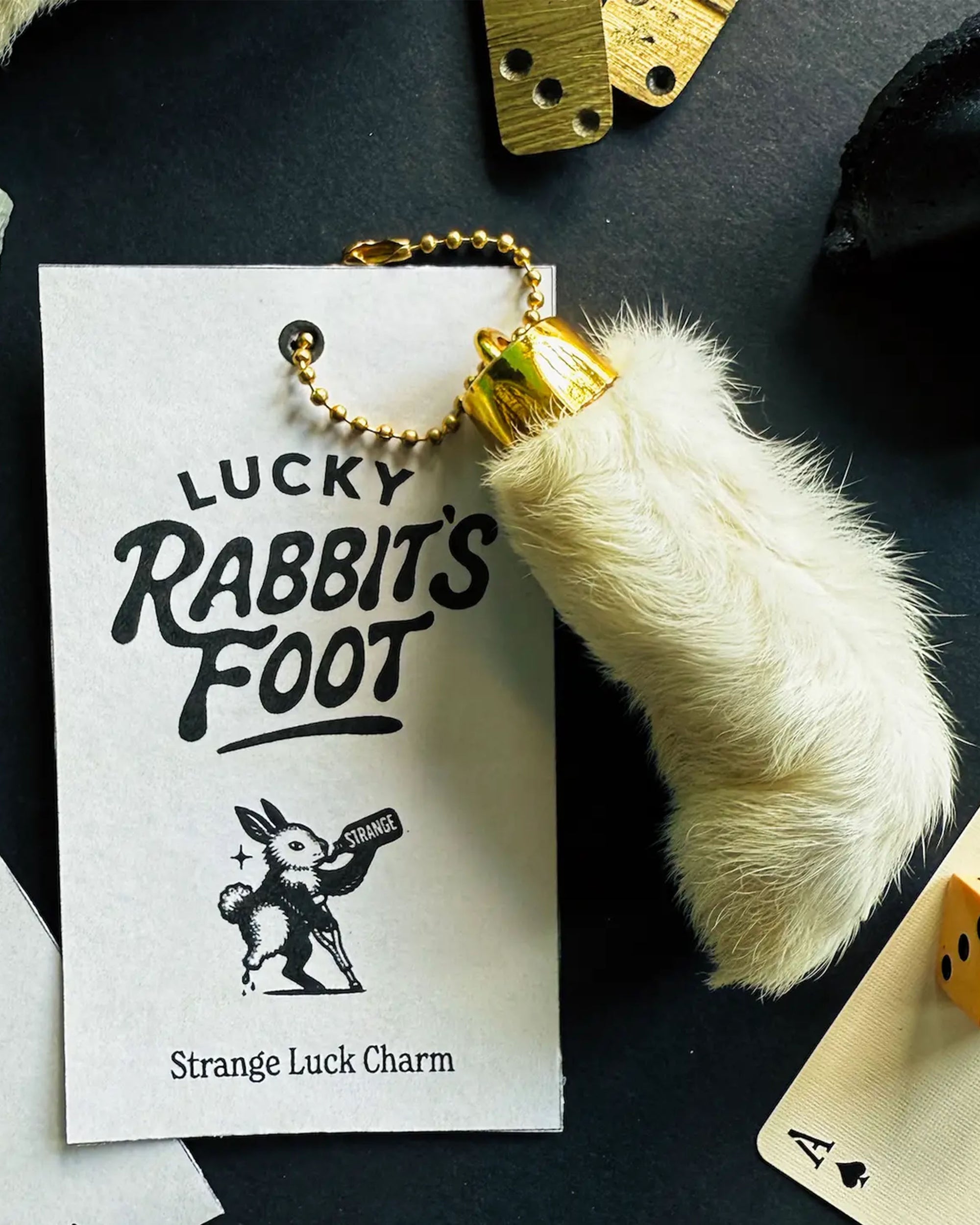The Faire Lucky Rabbit's Foot keychain features a white rabbit’s foot with a gold cap, attached to a card that reads “Lucky Rabbit’s Foot, Strange Luck Charm,” with playful artwork and surrounded by cards and dice.