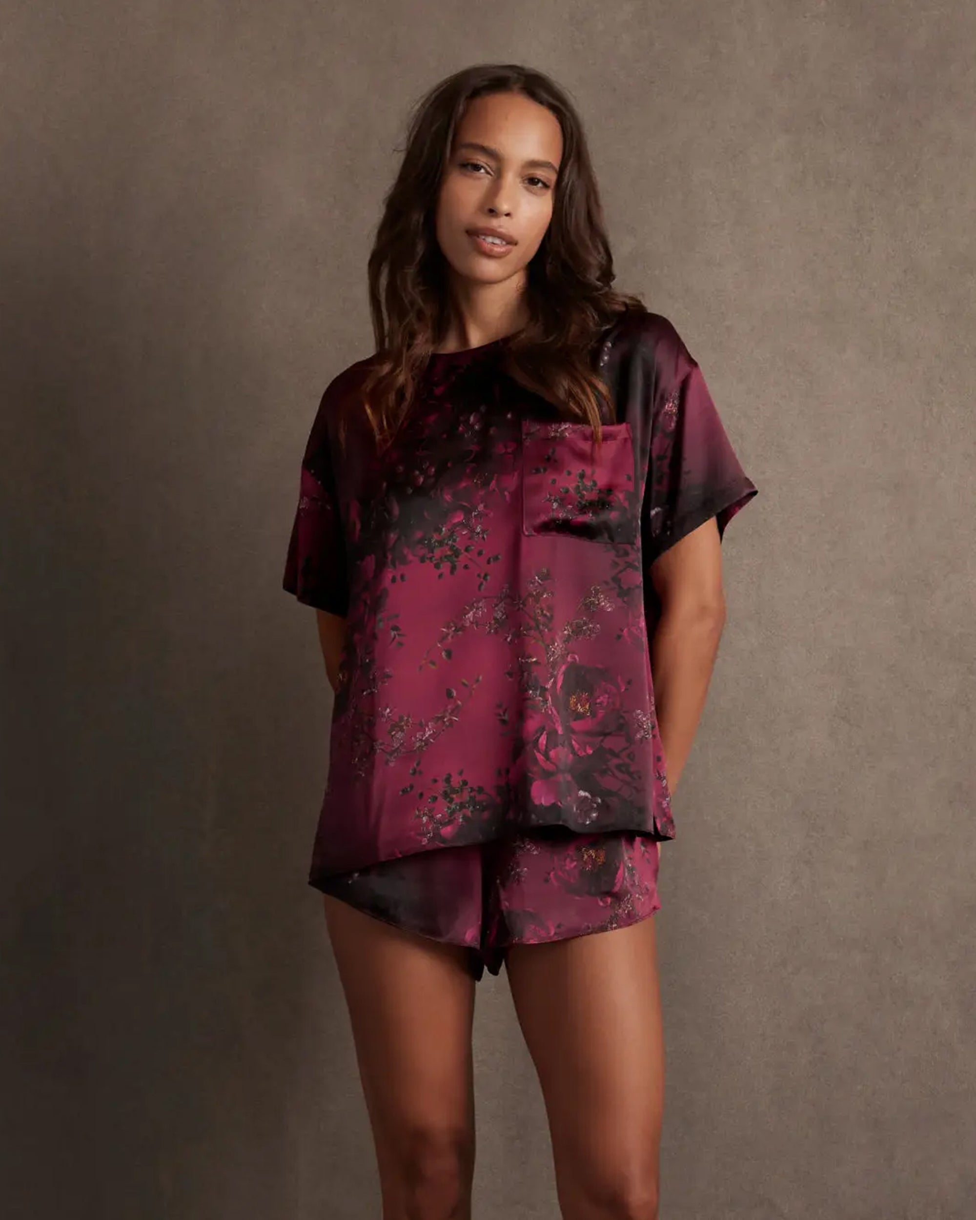 A woman stands against a plain background, wearing Faire's Lunya Washable Silk Tee and matching high-waist Lunya Washable Silk Shorts in deep burgundy with a subtle floral pattern.