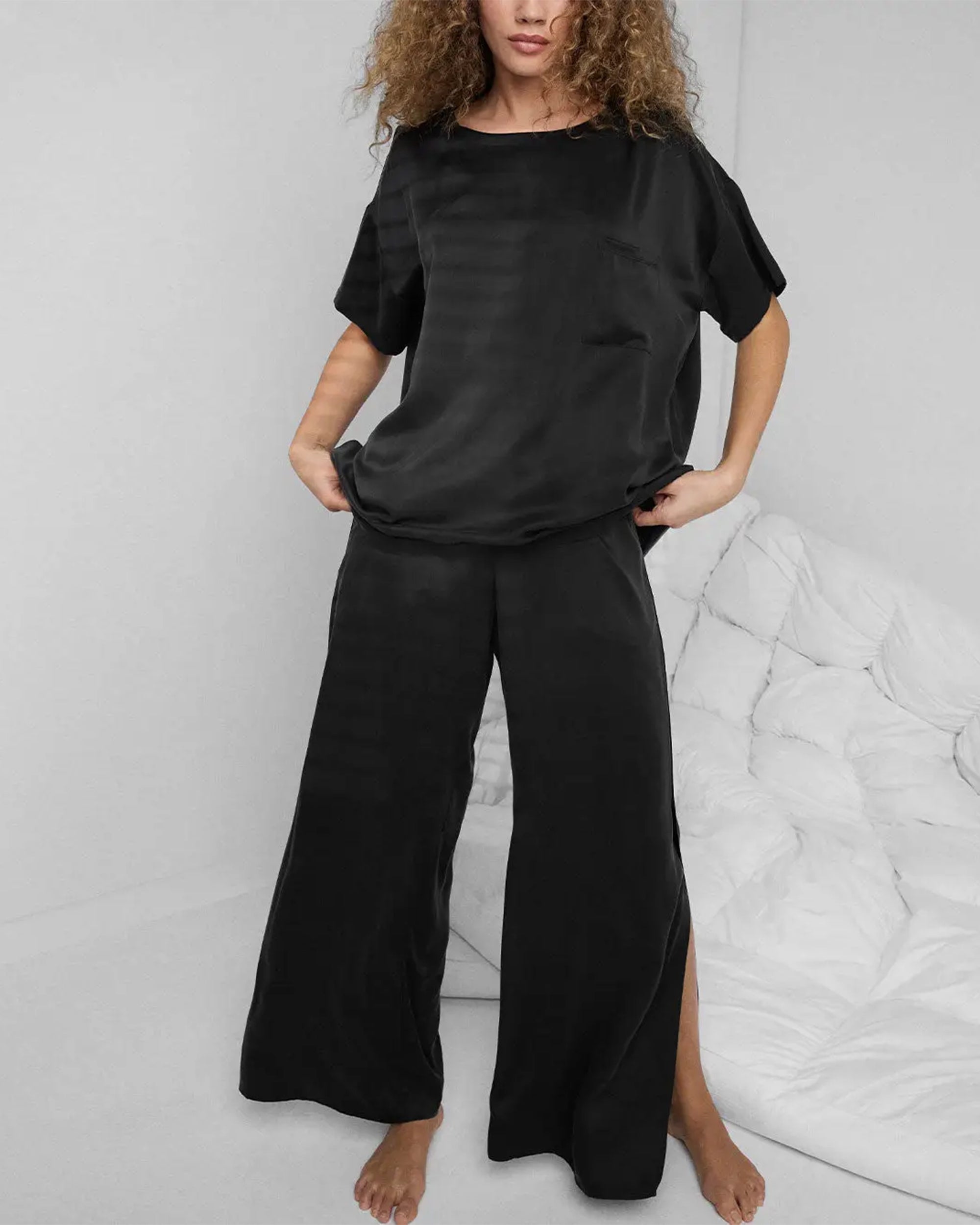 A woman with curly hair stands barefoot indoors, wearing Faire’s Lunya Washable Silk Pant and a matching silk top, posing casually beside a white tufted couch or bed.