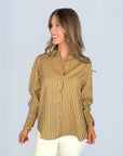 A woman with long, wavy hair smiles to the side, wearing the SUNCOO Lynsha Blouse—a brown and beige vertically striped button-down top with a collar—paired with off-white pants against a plain light background.
