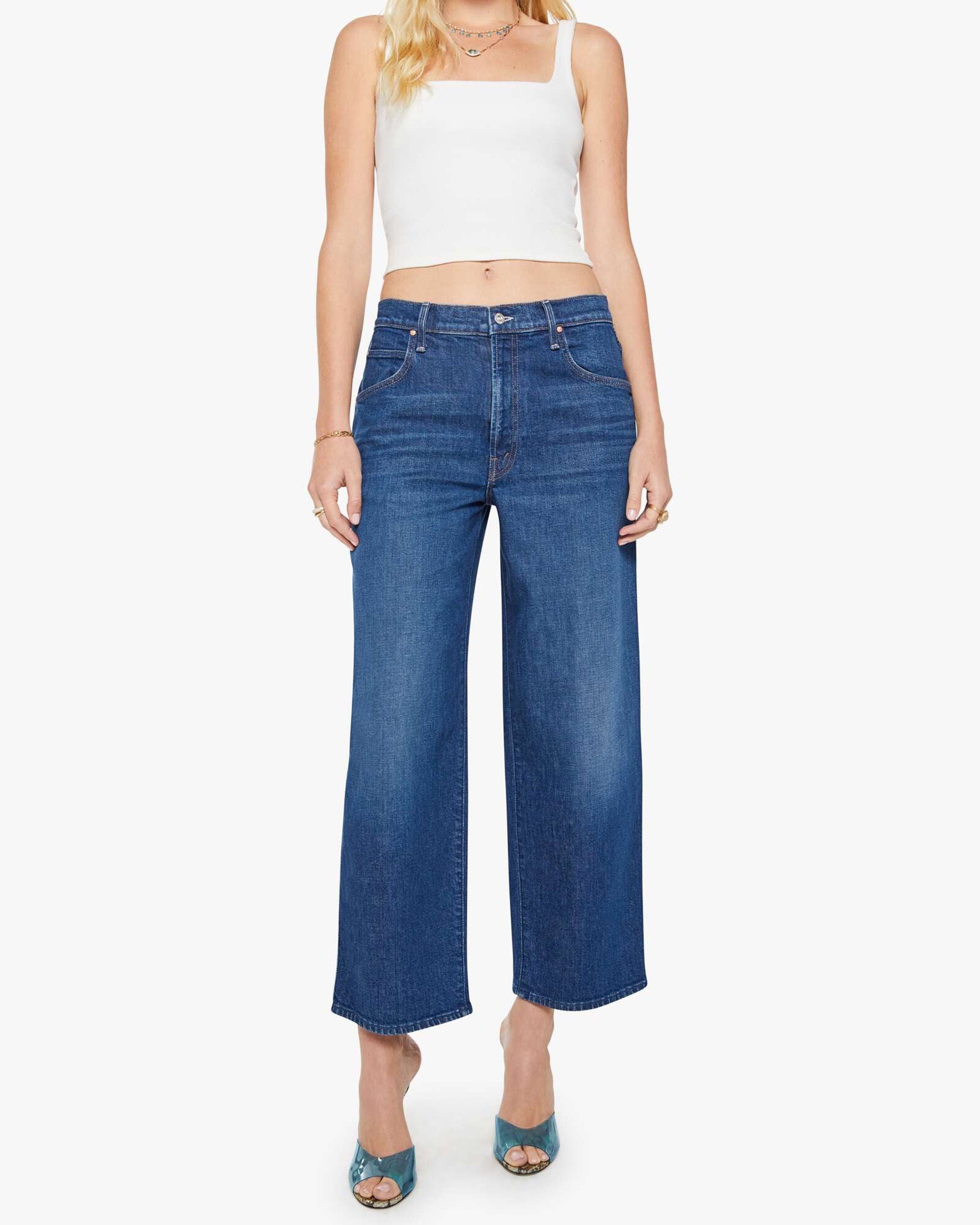 A woman wears The Delinquent Ankle dark blue oversized jeans by Mother, paired with a white sleeveless crop top and teal heeled sandals, standing against a plain white background.