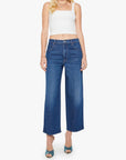 A woman wears The Delinquent Ankle dark blue oversized jeans by Mother, paired with a white sleeveless crop top and teal heeled sandals, standing against a plain white background.
