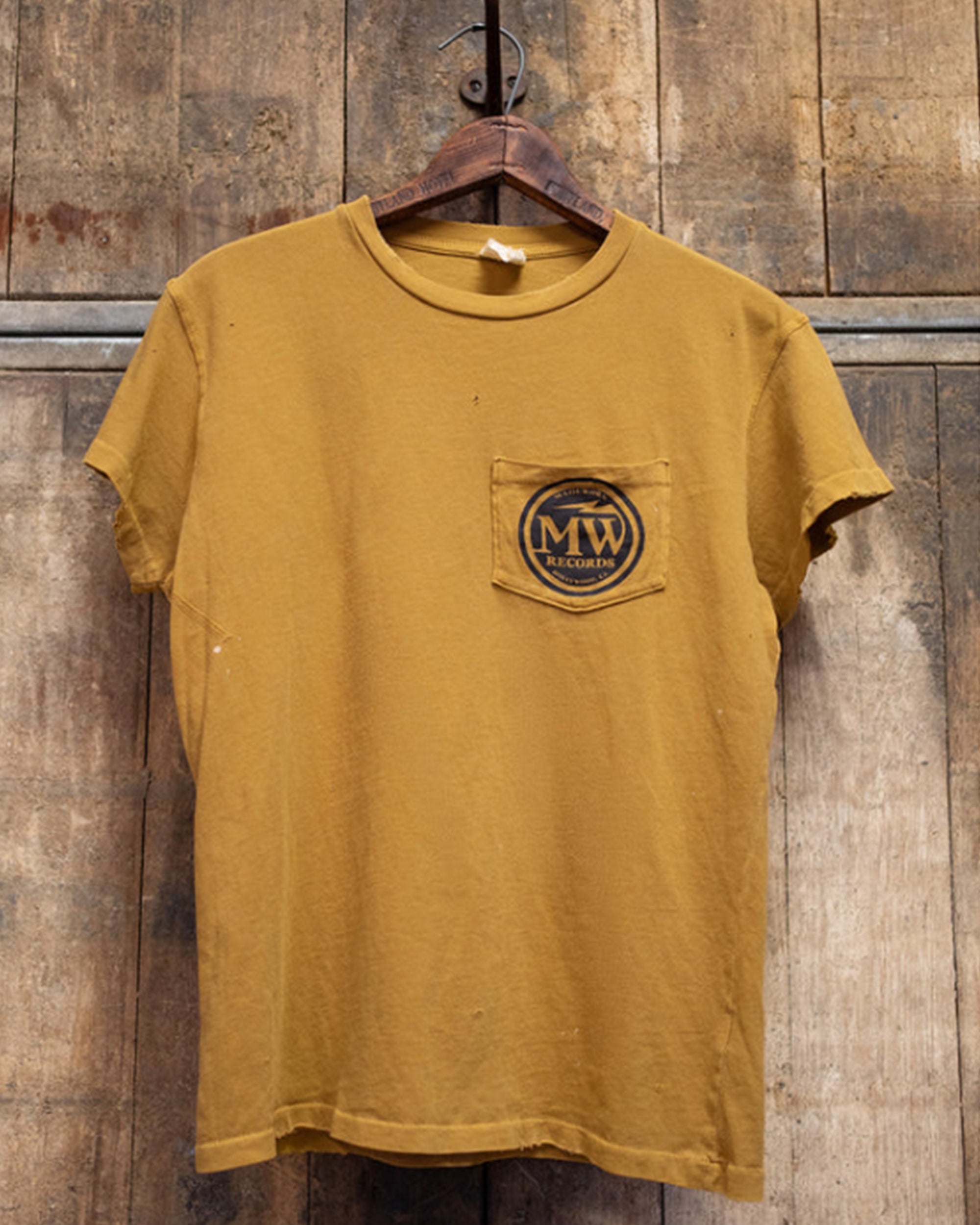 The Made Worn "MadeWorn Records" vintage unisex pocket tee in mustard yellow distressed cotton features a circular logo on the chest pocket. Handmade in Los Angeles, it’s shown on a wooden hanger with a rustic wood background.