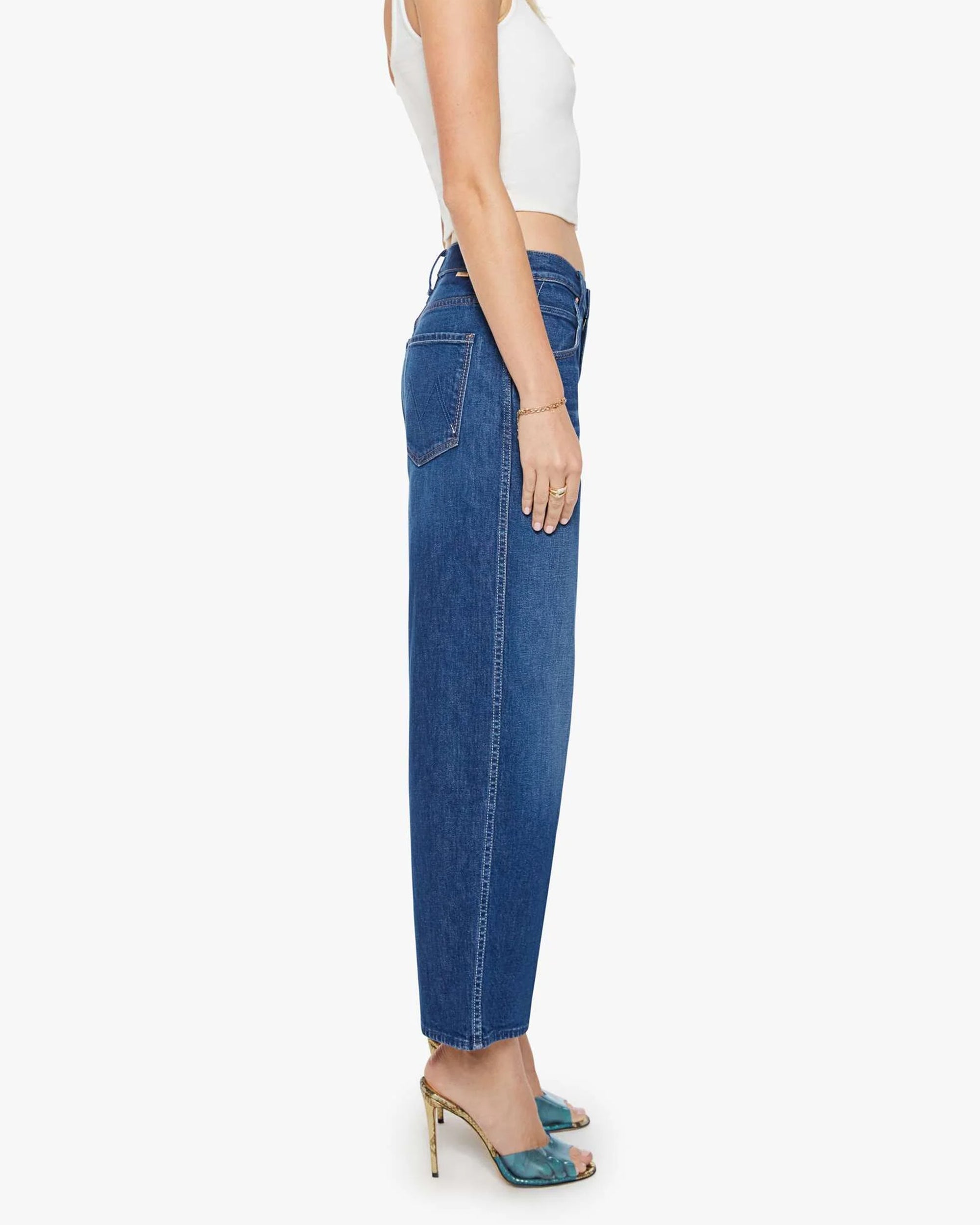 Woman stands sideways against a white background, wearing a white crop top, Mother’s The Delinquent Ankle high-waisted denim jeans, teal open-toe heeled sandals, and a bracelet.