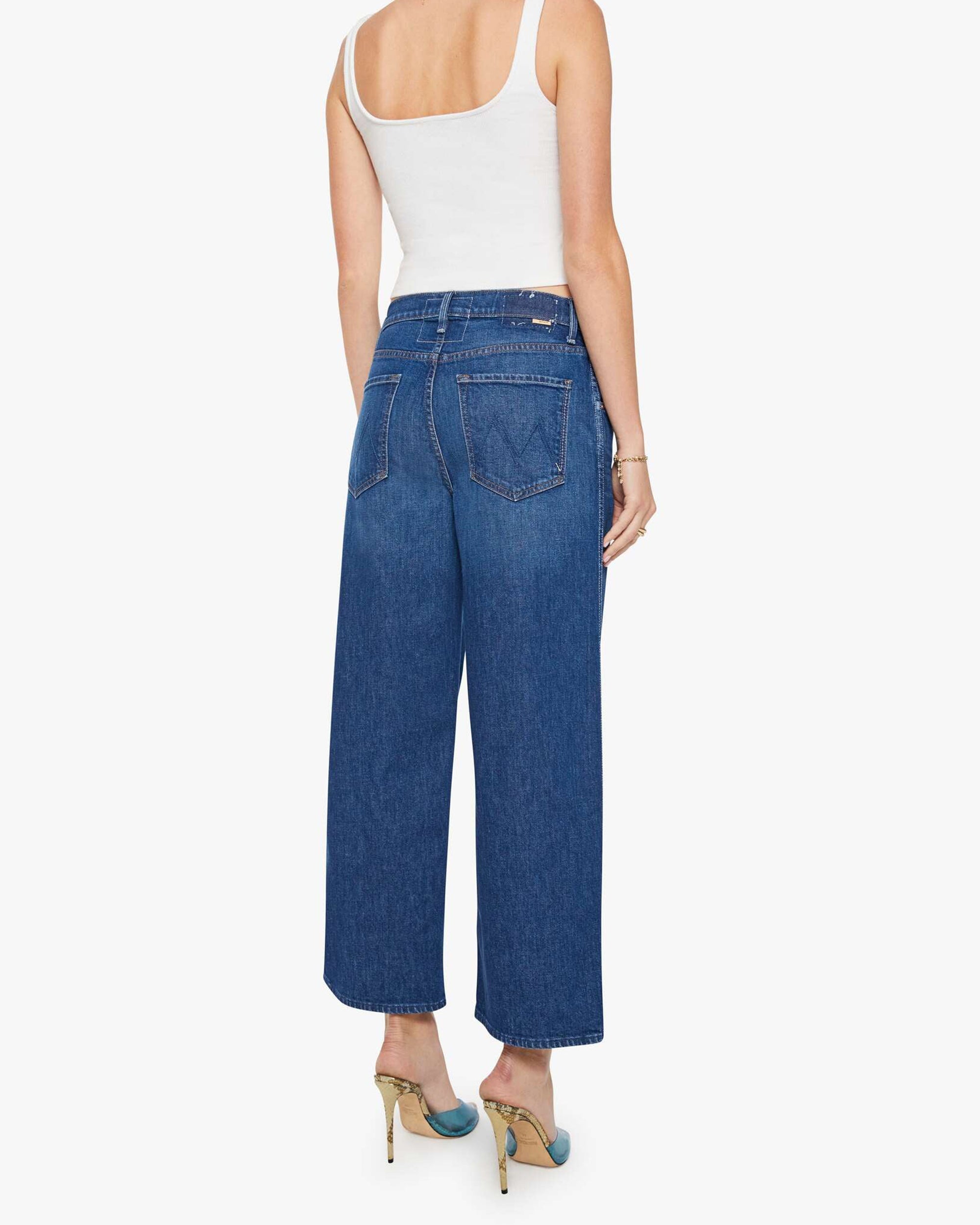 A woman faces away against a white background, wearing Mother's The Delinquent Ankle—ankle-length oversized jeans in premium denim—paired with a white tank top and green heels.