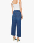 A woman faces away against a white background, wearing Mother's The Delinquent Ankle—ankle-length oversized jeans in premium denim—paired with a white tank top and green heels.