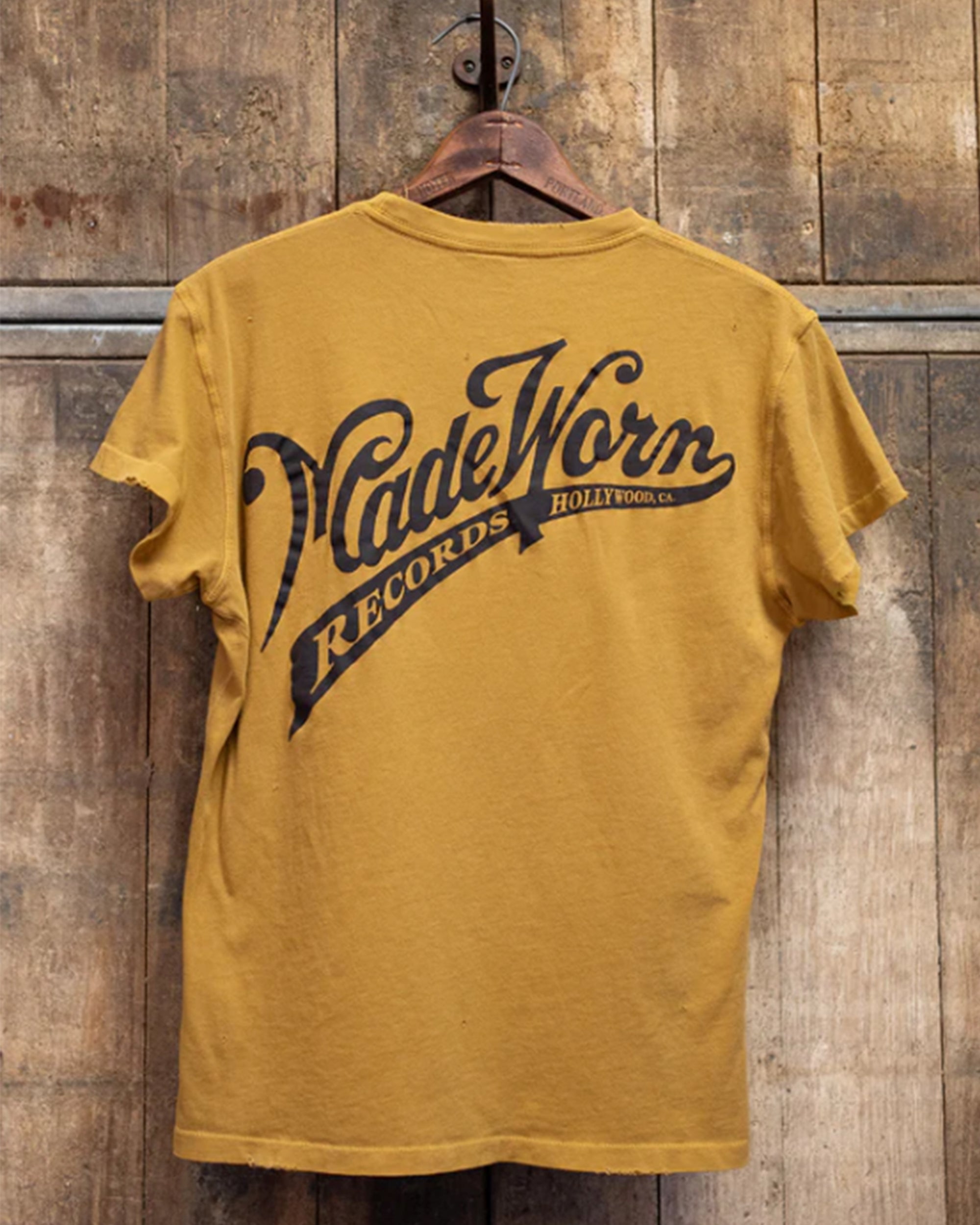 The MadeWorn Records unisex vintage pocket tee by Made Worn features mustard yellow distressed fabric and "RECORDS HOLLYWOOD, CA." text on the back. It hangs on a wooden hanger against a rustic wall and is made in Los Angeles.