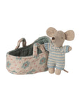 The Maileg Baby Mouse in Carry Cot by Maileg features a tiny mouse in a blue and white knitted suit, standing beside a floral-patterned fabric carry cot with two handles.