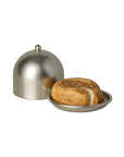 The Maileg Cheese Bell by Maileg features a realistic bagel-shaped treat on a small round metal dish, with a matching dome cover beside it, perfect as a mouse snack and displayed against a white background.