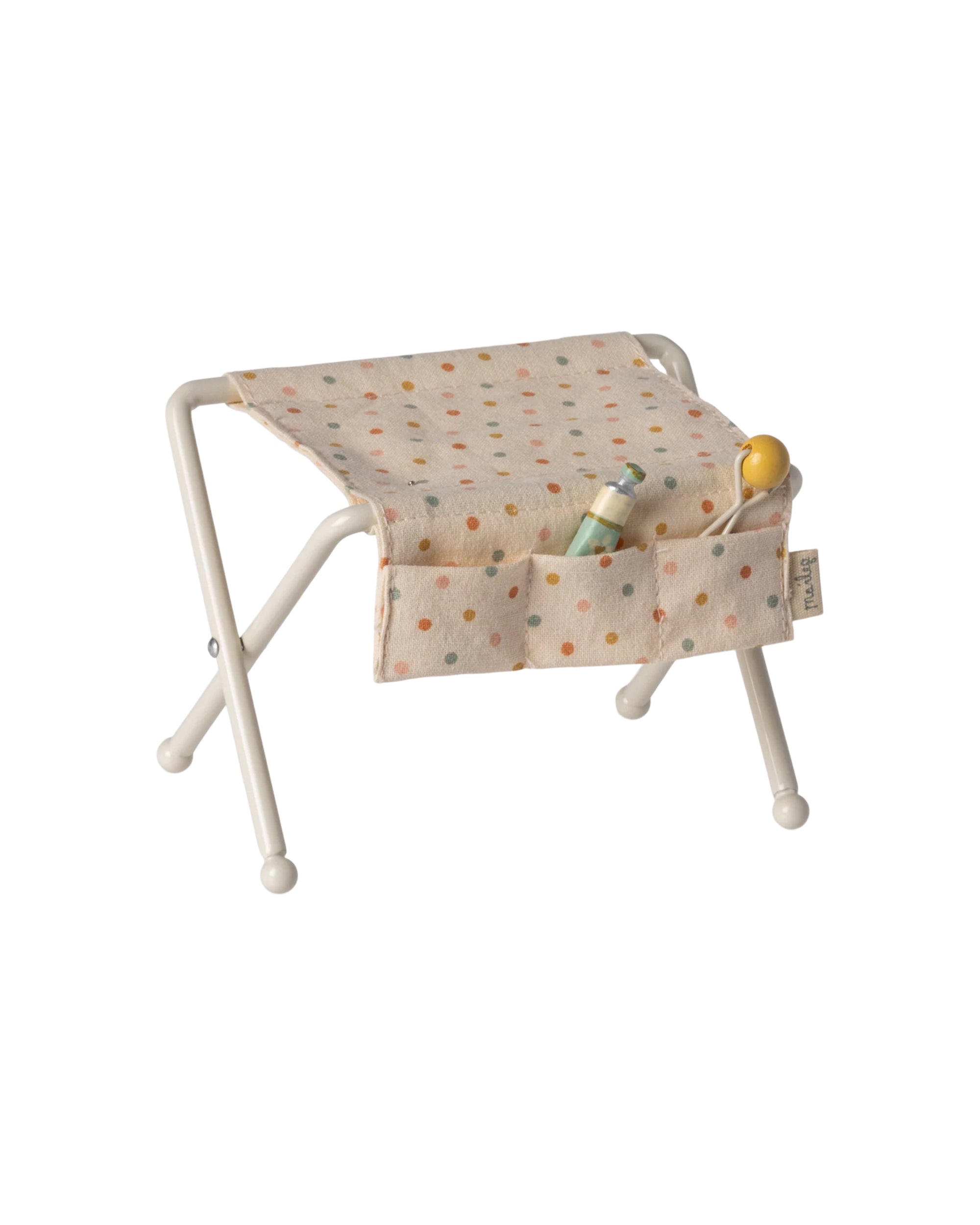 The Maileg Nursery Table by Maileg is a small white metal folding stool with a beige polka dot seat and front pockets for baby accessories, ideal for a nursery or tiny baby mice to use.