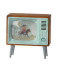 The Maileg Television by Maileg is a small, retro-style TV with a wood frame and metal legs, featuring a cartoon cowboy on horseback with a cactus in the background displayed on its screen.