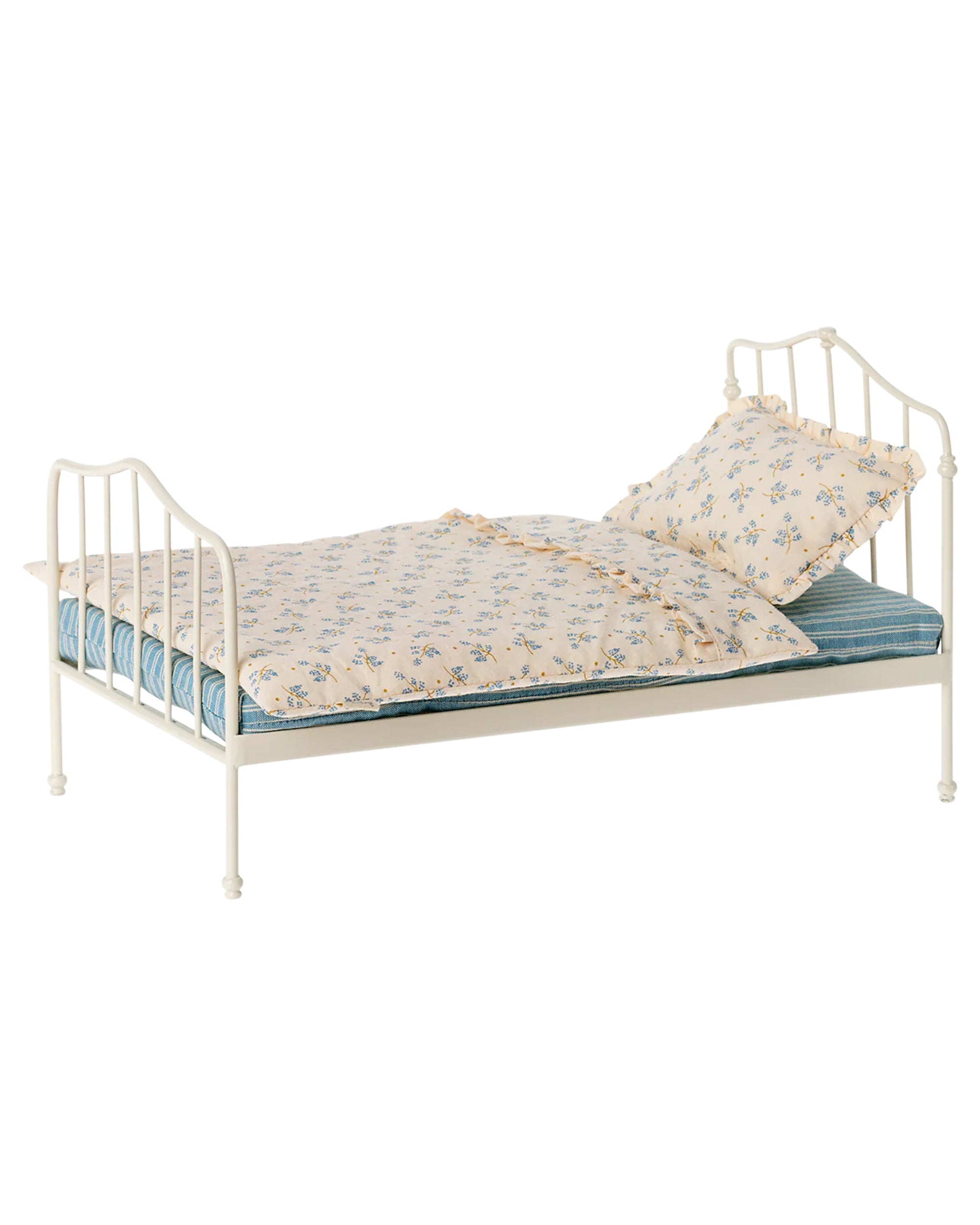 The Maileg Vintage Bed, Miniature by Maileg features a small white metal frame with a vintage design, blue-striped mattress, cream blanket, floral pillow, and decorative headboard and footboard with vertical bars.
