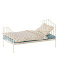 The Maileg Vintage Bed, Miniature by Maileg features a small white metal frame with a vintage design, blue-striped mattress, cream blanket, floral pillow, and decorative headboard and footboard with vertical bars.