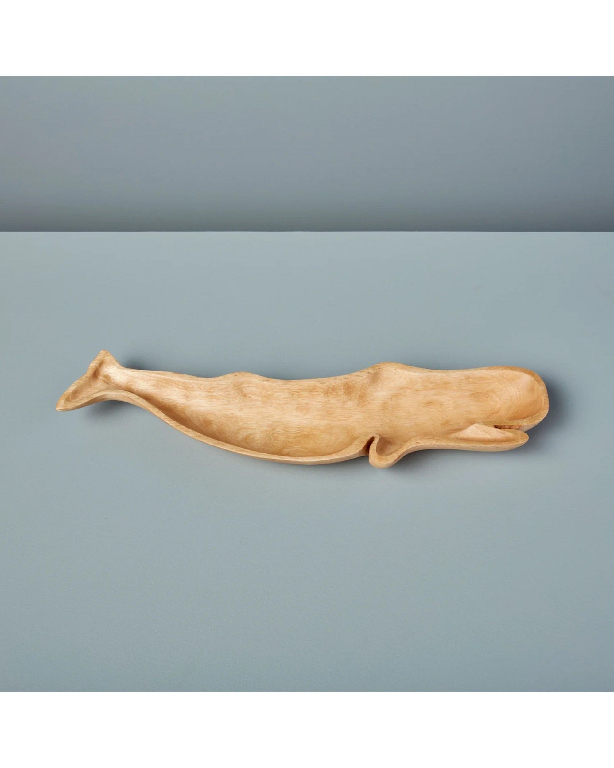 The Be Home Kiln Mango Wood Whale Dish, handcarved from light brown mango wood, is shown on a smooth light blue surface with a plain blue background.