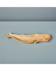 The Be Home Kiln Mango Wood Whale Dish, handcarved from light brown mango wood, is shown on a smooth light blue surface with a plain blue background.