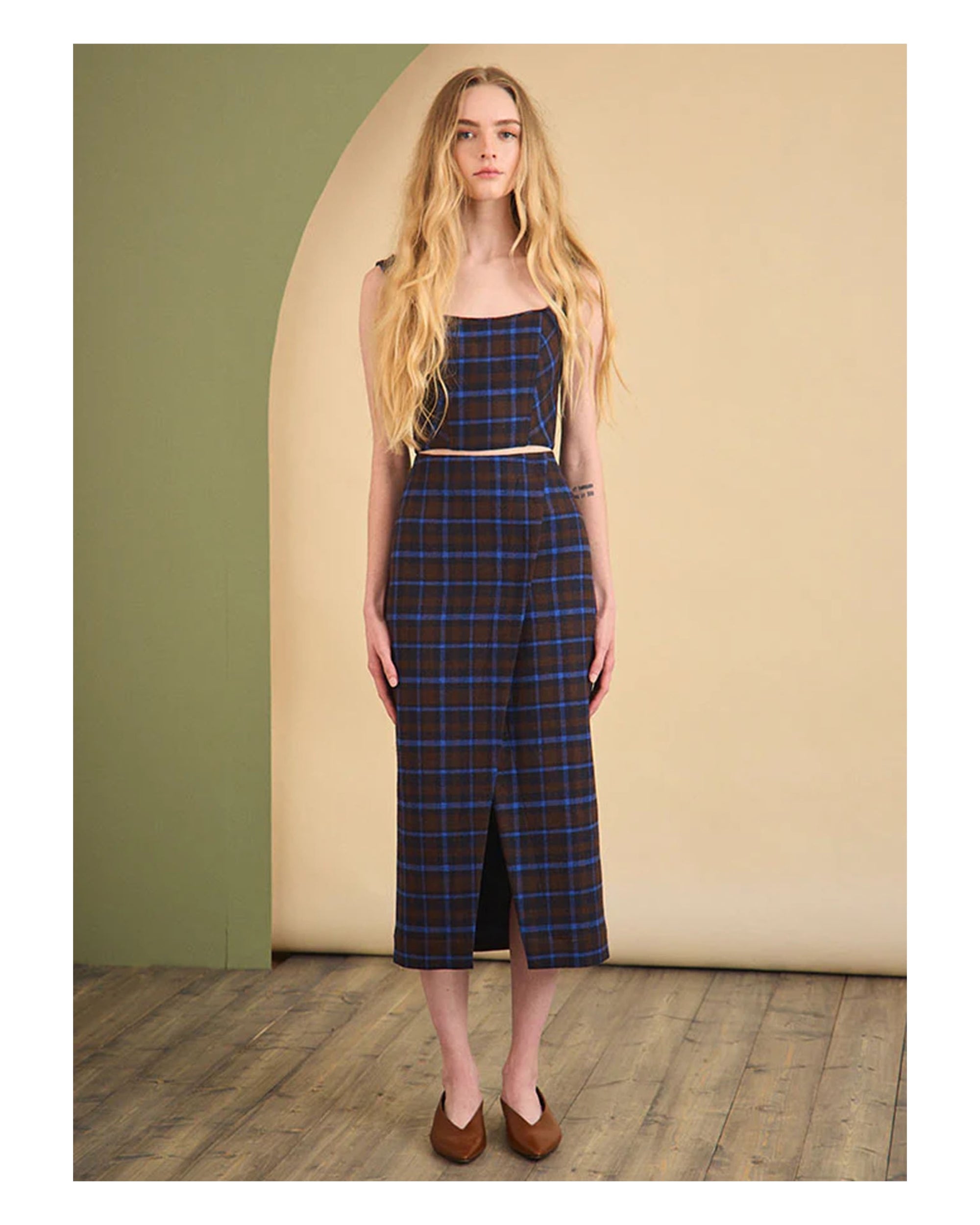 A woman with long blonde hair wears the Hunter Bell NYC Manor Wrap Skirt, a sleeveless, square-neck dress with an Aspen Plaid skirt and front slit, standing on a wooden floor against a green and beige background.