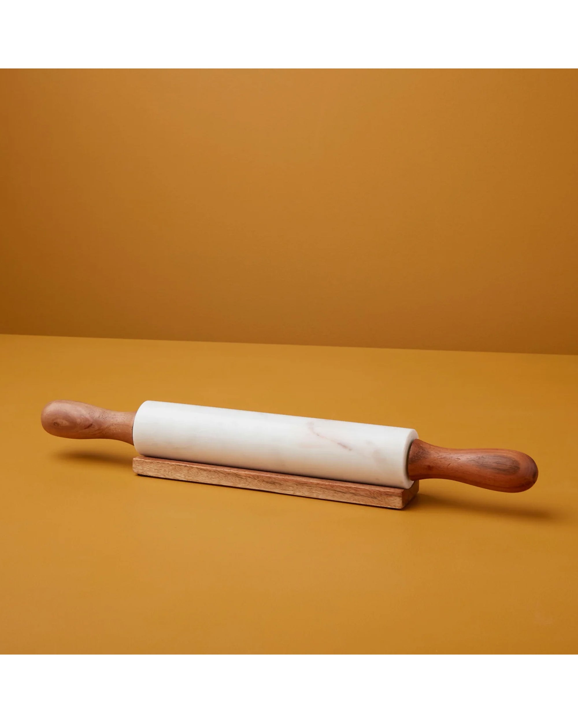 The Be Home Rolling White Marble Acacia Pin features marble with wooden handles and a wooden base, displayed against a solid mustard yellow background.