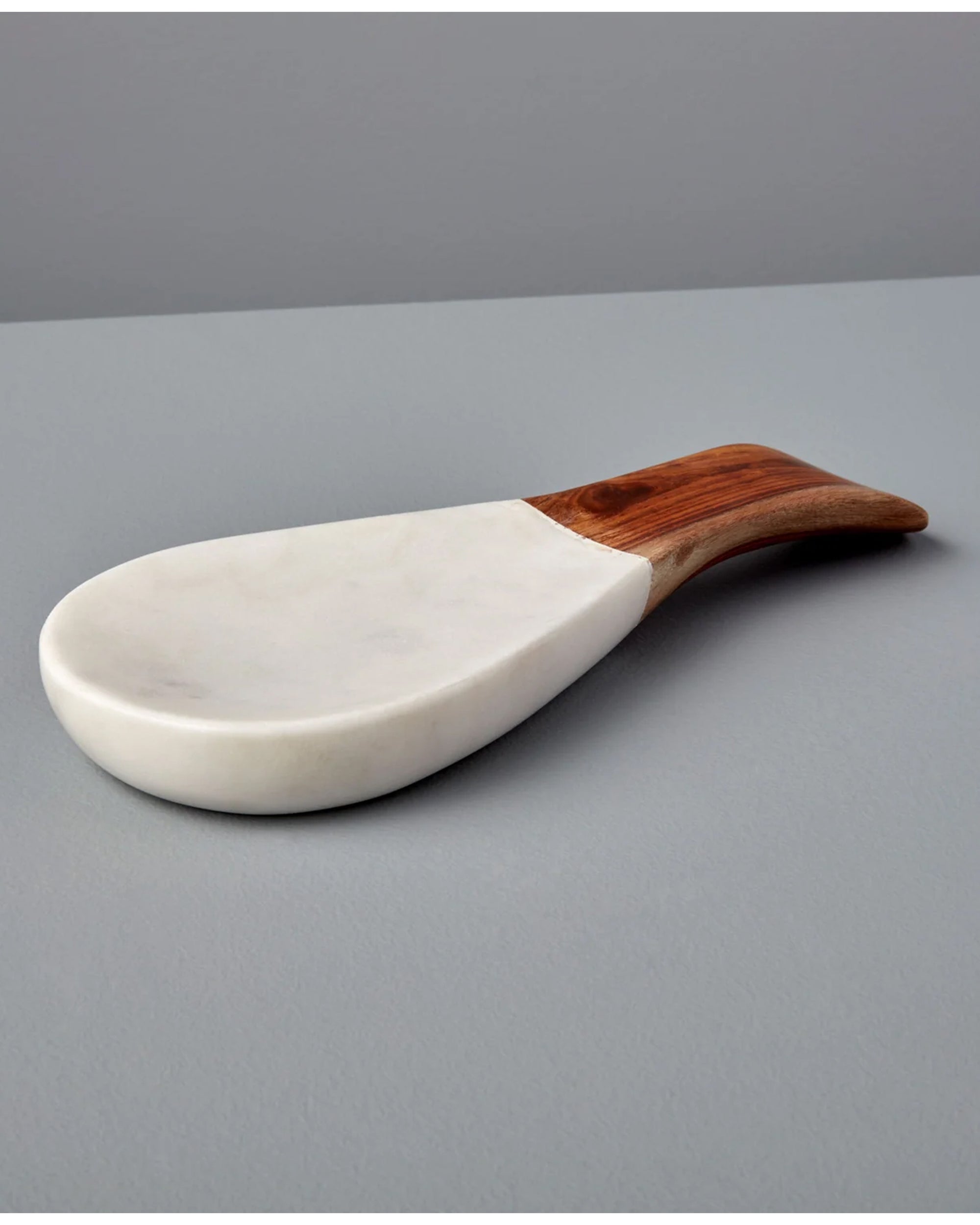 The Be Home White Marble Wood Spoon Rest features a wide, flat white marble base and a handcrafted acacia wood handle, elegantly placed on a smooth gray surface.