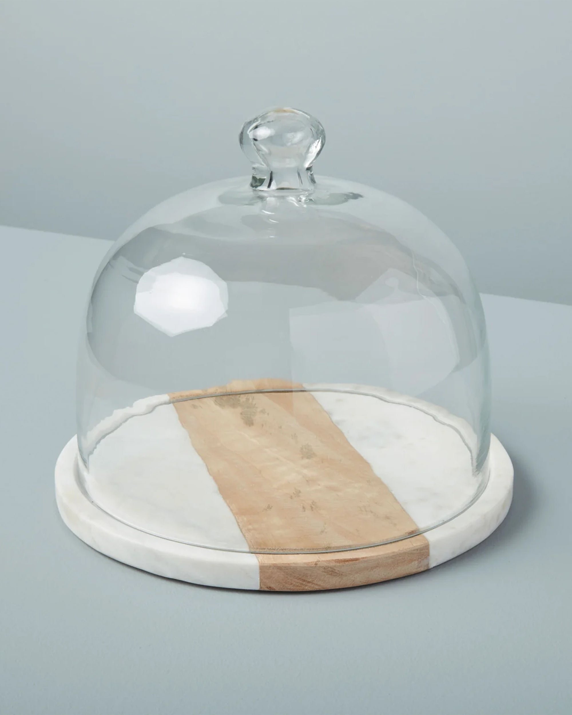 The Be Home White Marble Wood Glass Cloche features a glass dome with a knob handle on a round board of white marble and handcrafted acacia, part of the Pavia Collection, shown against a light grey background.