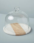The Be Home White Marble Wood Glass Cloche features a glass dome with a knob handle on a round board of white marble and handcrafted acacia, part of the Pavia Collection, shown against a light grey background.