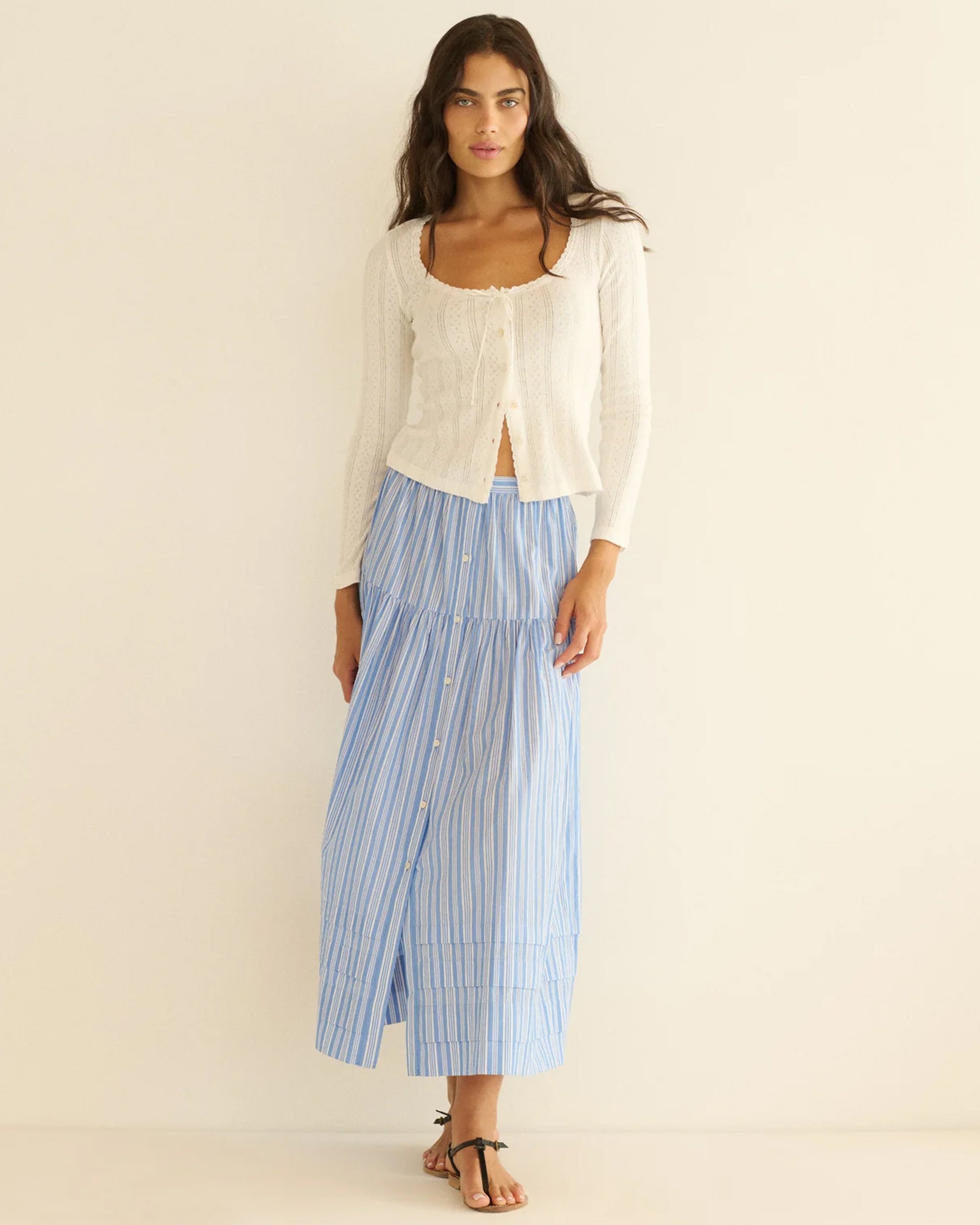 A woman with long dark hair and a relaxed expression stands against a plain light wall, wearing a white long-sleeve cardigan, the Marceline Skirt by DOEN—a striped tiered organic cotton piece—and black flat sandals.