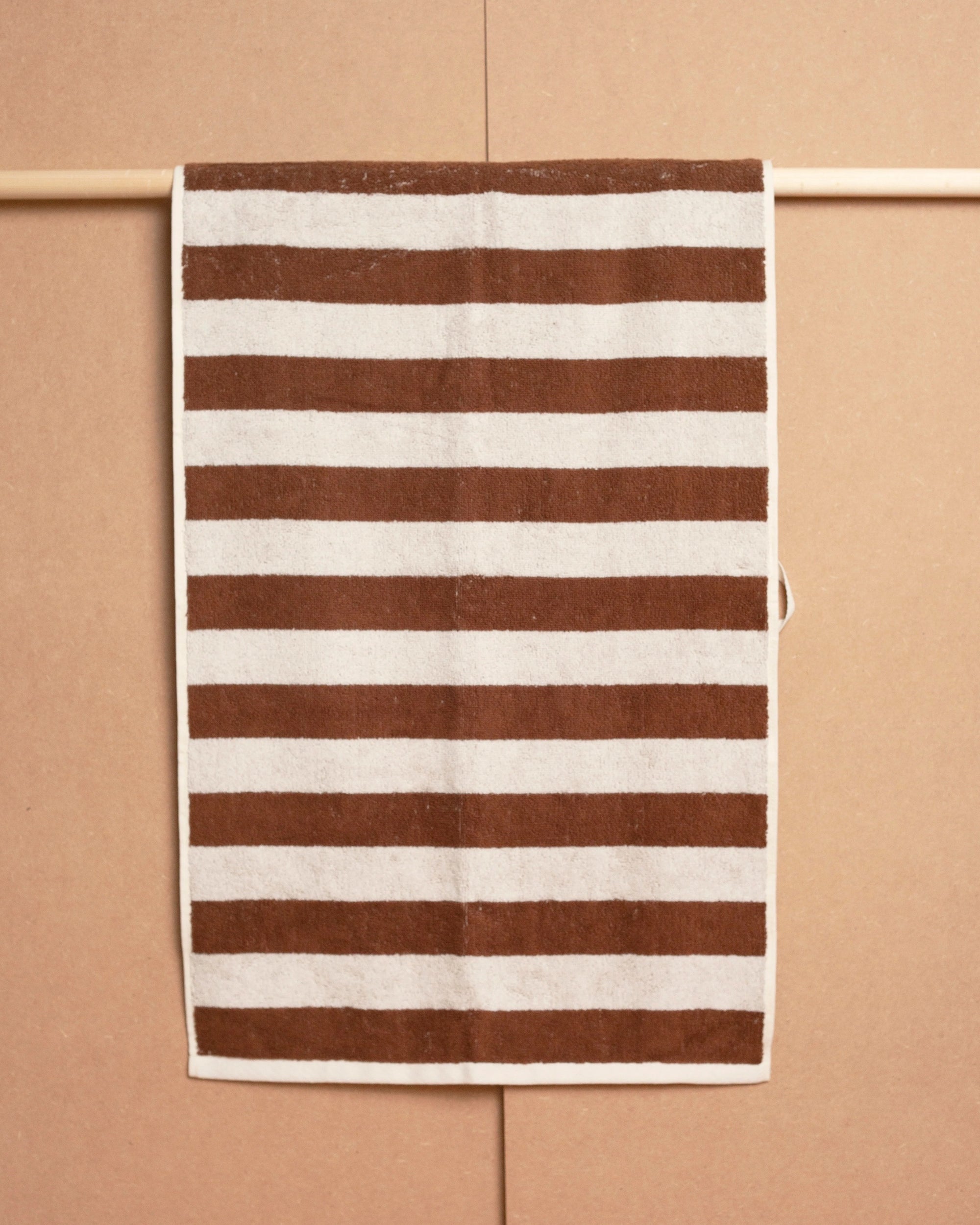 The Maria Hand Towel by Autumn Sonata, featuring a jacquard weave and brown and white horizontal stripes, is draped neatly over a wooden rod against a beige background.