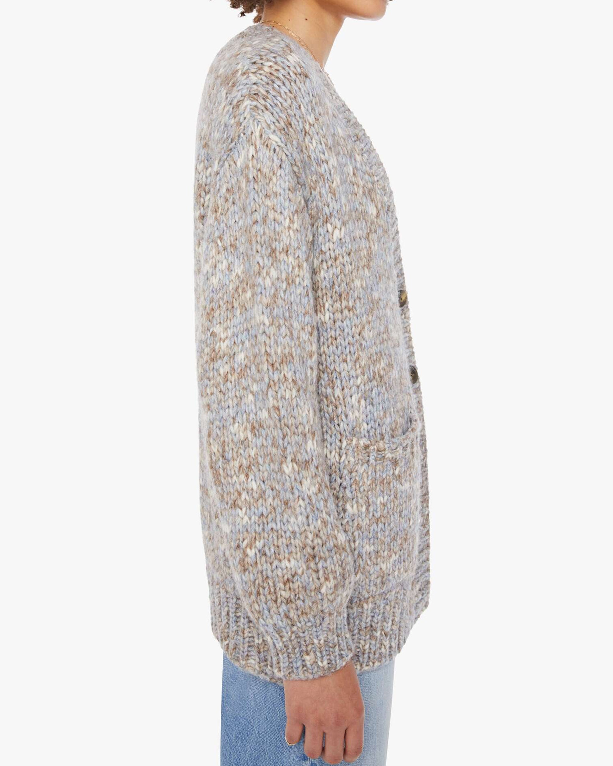 A person stands sideways in the Xirena Marvin Sweater—an oversized, chunky, multi-colored knit cardigan with large buttons and front pockets—paired with light blue jeans against a plain white background.