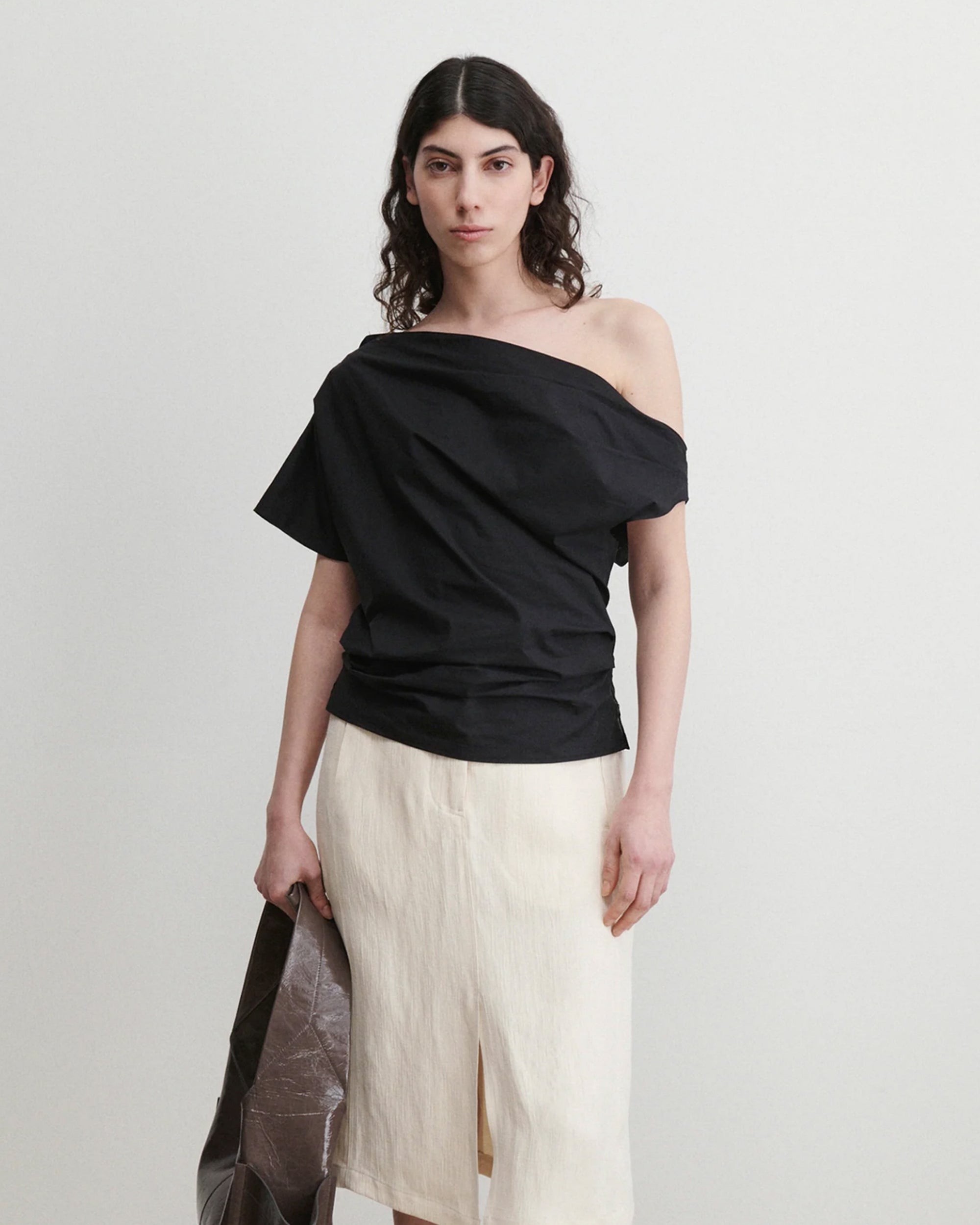 A person with long, dark, wavy hair wears the Rachel Comey Mata Top in black and a cream organic cotton skirt with a front slit, holding a large brown bag against a plain light background.