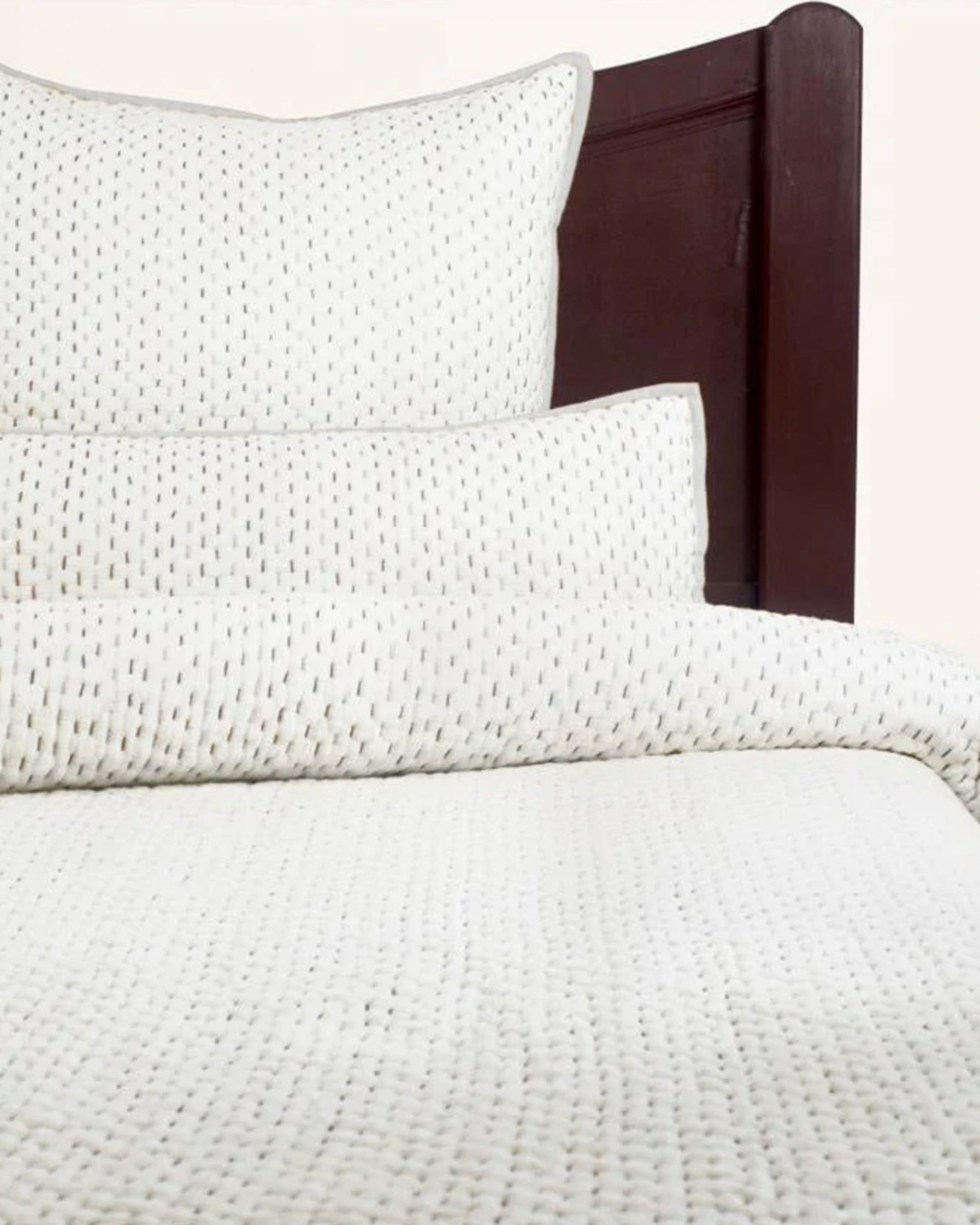 A close-up of a neatly made bed showcases the Avasa Bedding Maya Quilt Shades of Cream Queen, featuring hand-quilted texture and artisanal stitching, set against a dark wooden headboard.
