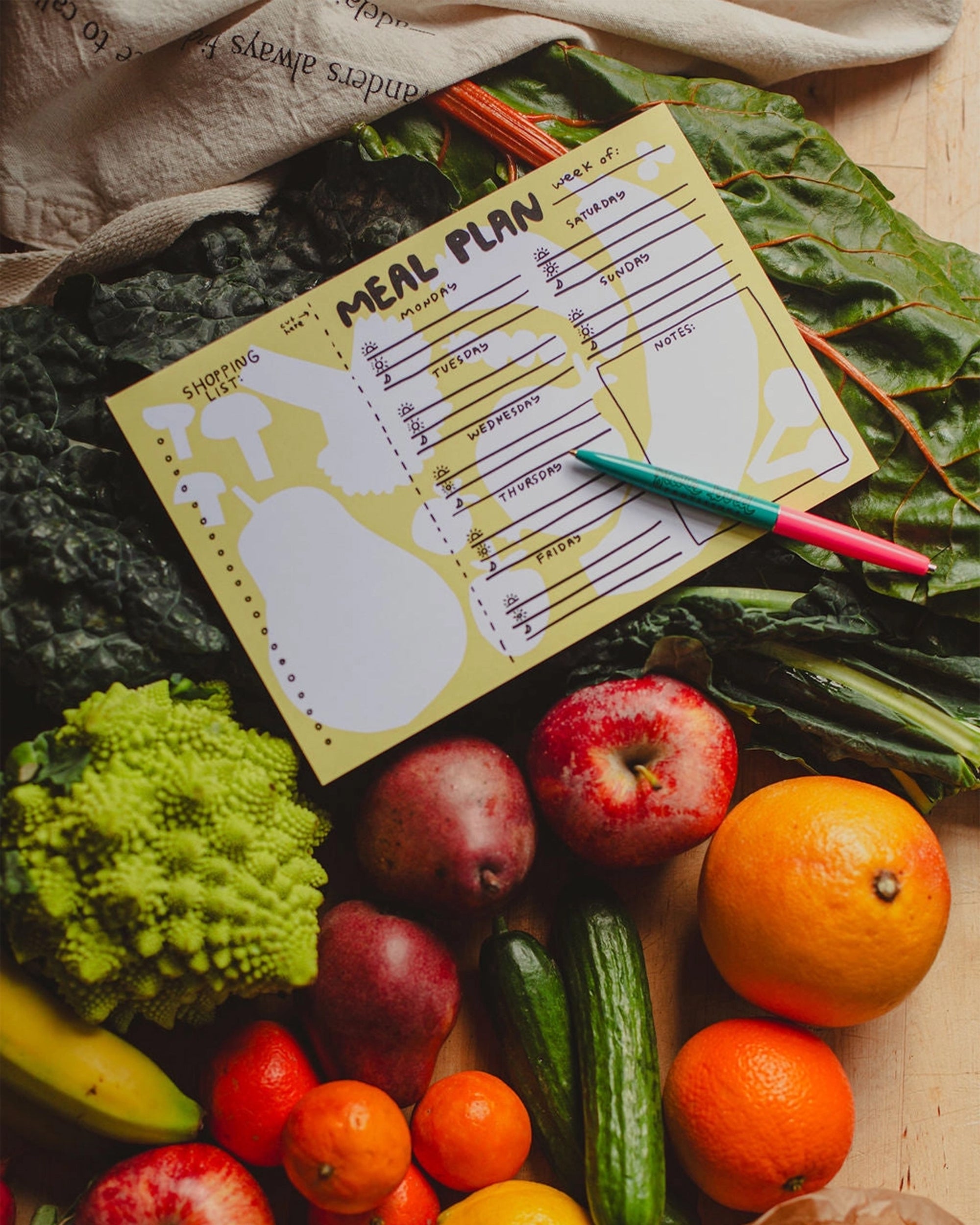 The Faire Meal Planner Notepad, proudly made in the USA, features handwritten days and meals. Shown atop leafy greens with fresh fruits and veggies—like oranges, apples, bananas, cucumbers, and broccoli—inspires healthy planning.