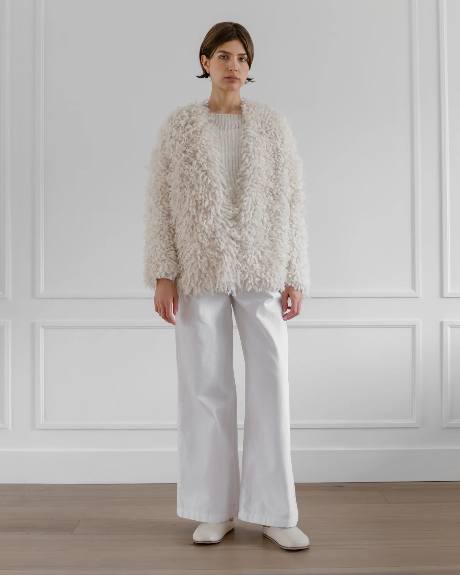 A person stands indoors against white paneled walls and a wooden floor, wearing LATIERRA's handcrafted Medusa Cardigan Coat—white and fluffy—paired with white wide-leg pants, a white top, and white shoes.