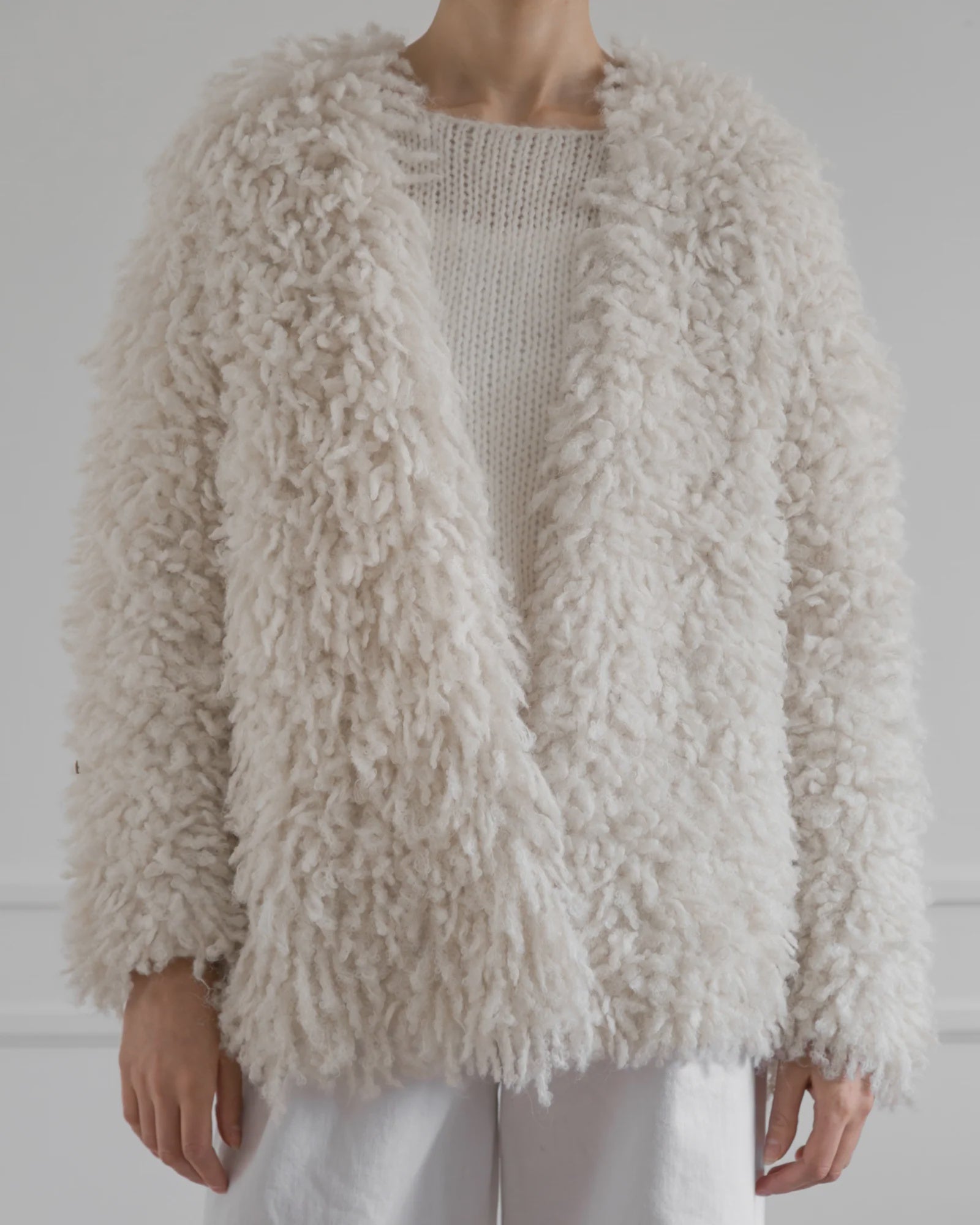 A person wearing the cream Medusa Cardigan Coat by LATIERRA over a knitted sweater and white pants stands against a light background—embodying sustainable luxury and cozy style.