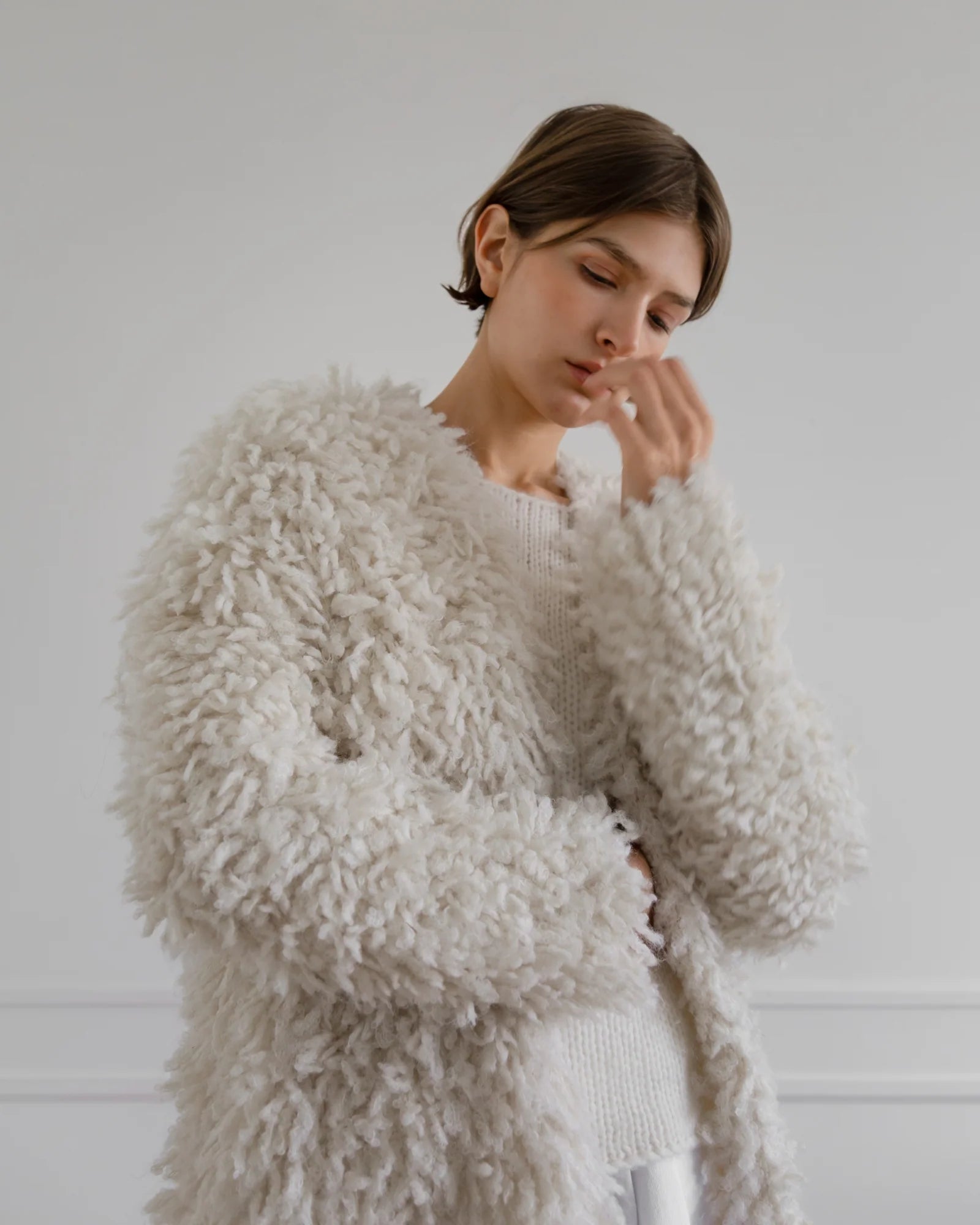 A person with short brown hair wears the LATIERRA Medusa Cardigan Coat, an off-white textured piece, layered over a white sweater, standing against a light gray background and gently touching their face.