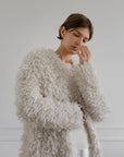 A person with short brown hair wears the LATIERRA Medusa Cardigan Coat, an off-white textured piece, layered over a white sweater, standing against a light gray background and gently touching their face.