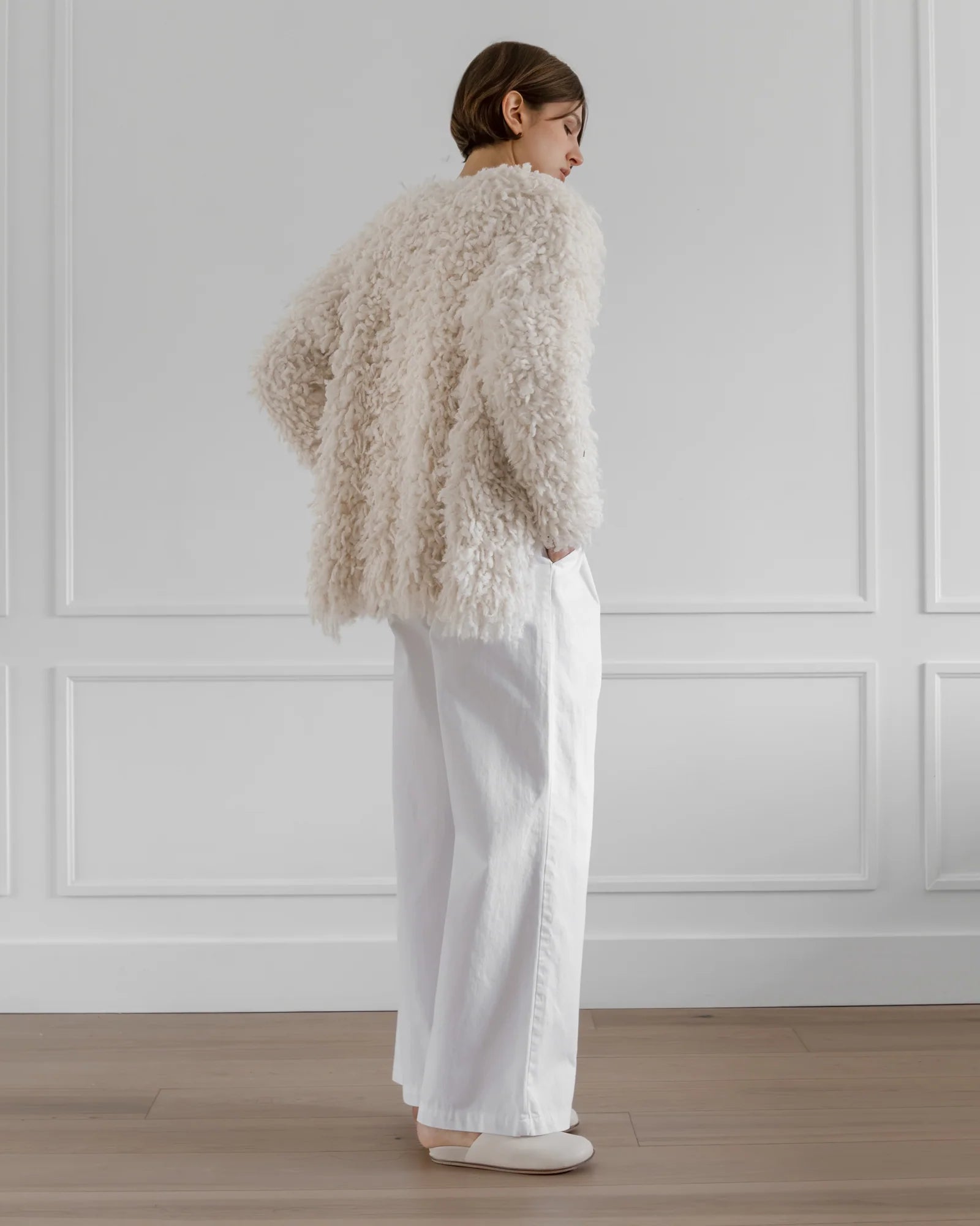 A person stands sideways in a minimalistic room, wearing the LATIERRA Medusa Cardigan Coat—an off-white handcrafted alpaca piece—paired with loose white pants and shoes. The space’s light wood floor and white, molded walls exude sustainable luxury.