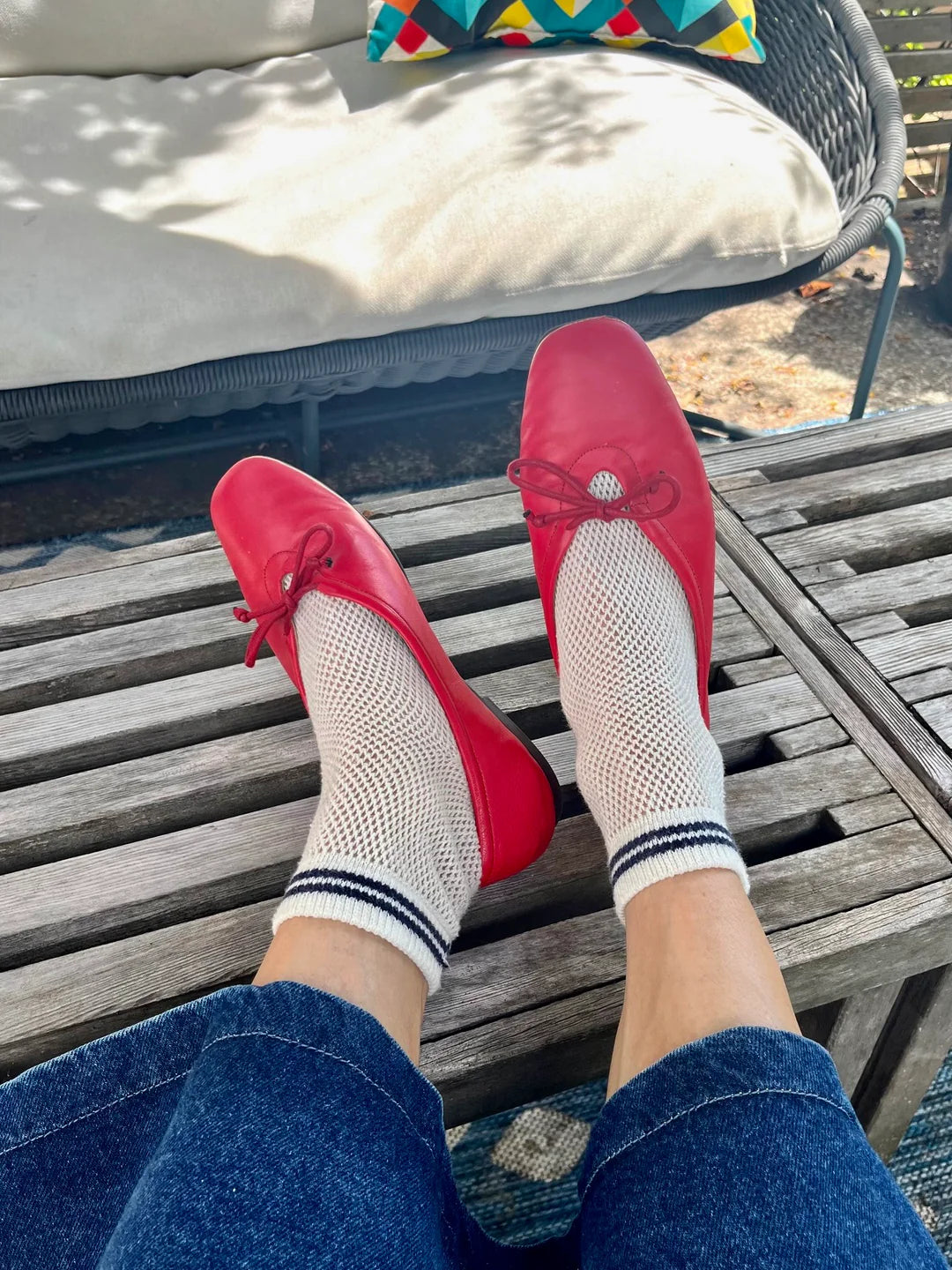A person wearing Unified Mesh Sneaker Socks with black stripes and red ballet flats rests their feet on a wooden slatted table, surrounded by wicker outdoor furniture and a colorful pillow for a retro sport vibe.