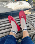 A person wearing Unified Mesh Sneaker Socks with black stripes and red ballet flats rests their feet on a wooden slatted table, surrounded by wicker outdoor furniture and a colorful pillow for a retro sport vibe.